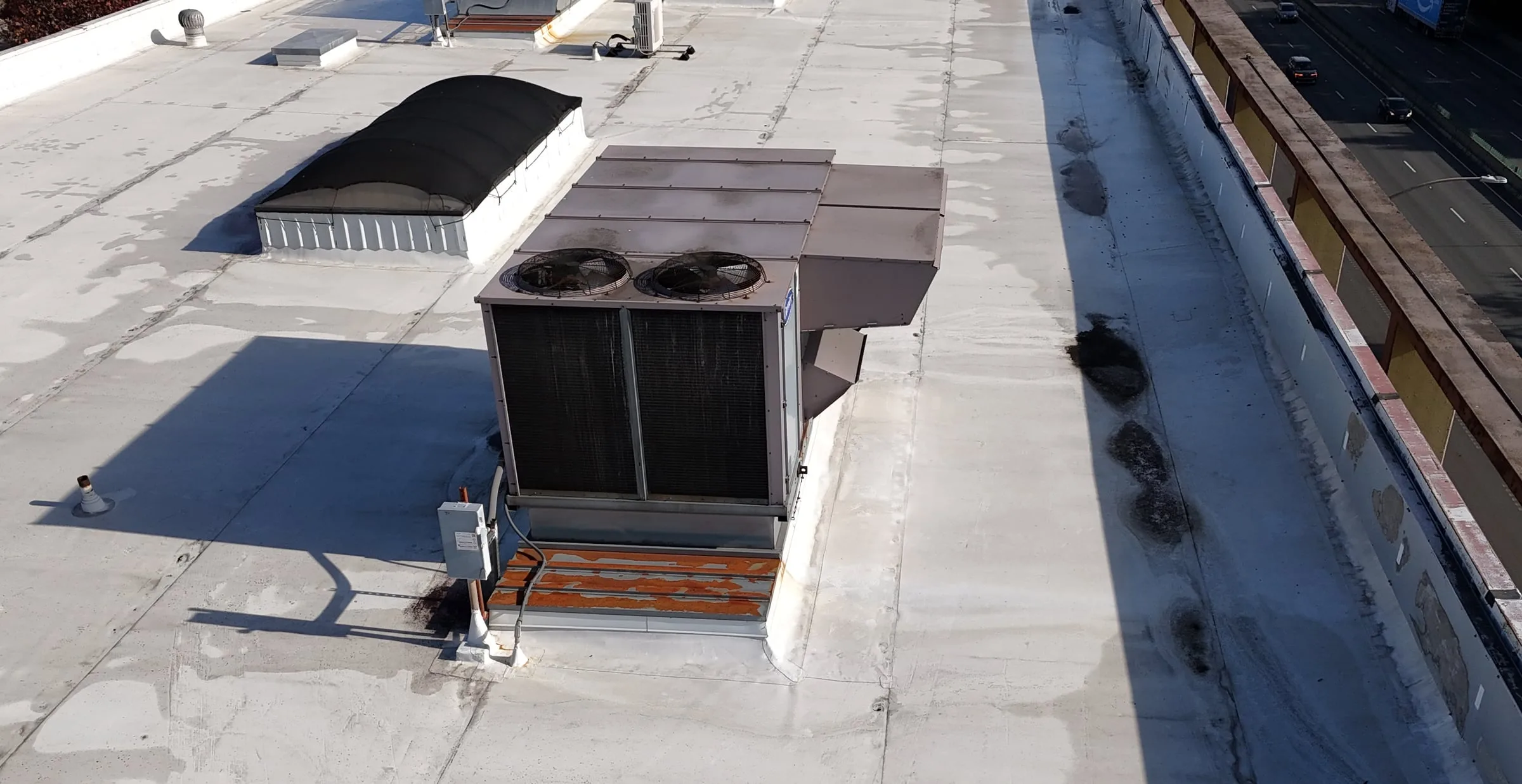 A large AC unit on a white flat roof, with the roofing wrapped up the sides of its curb and a power line running to it.