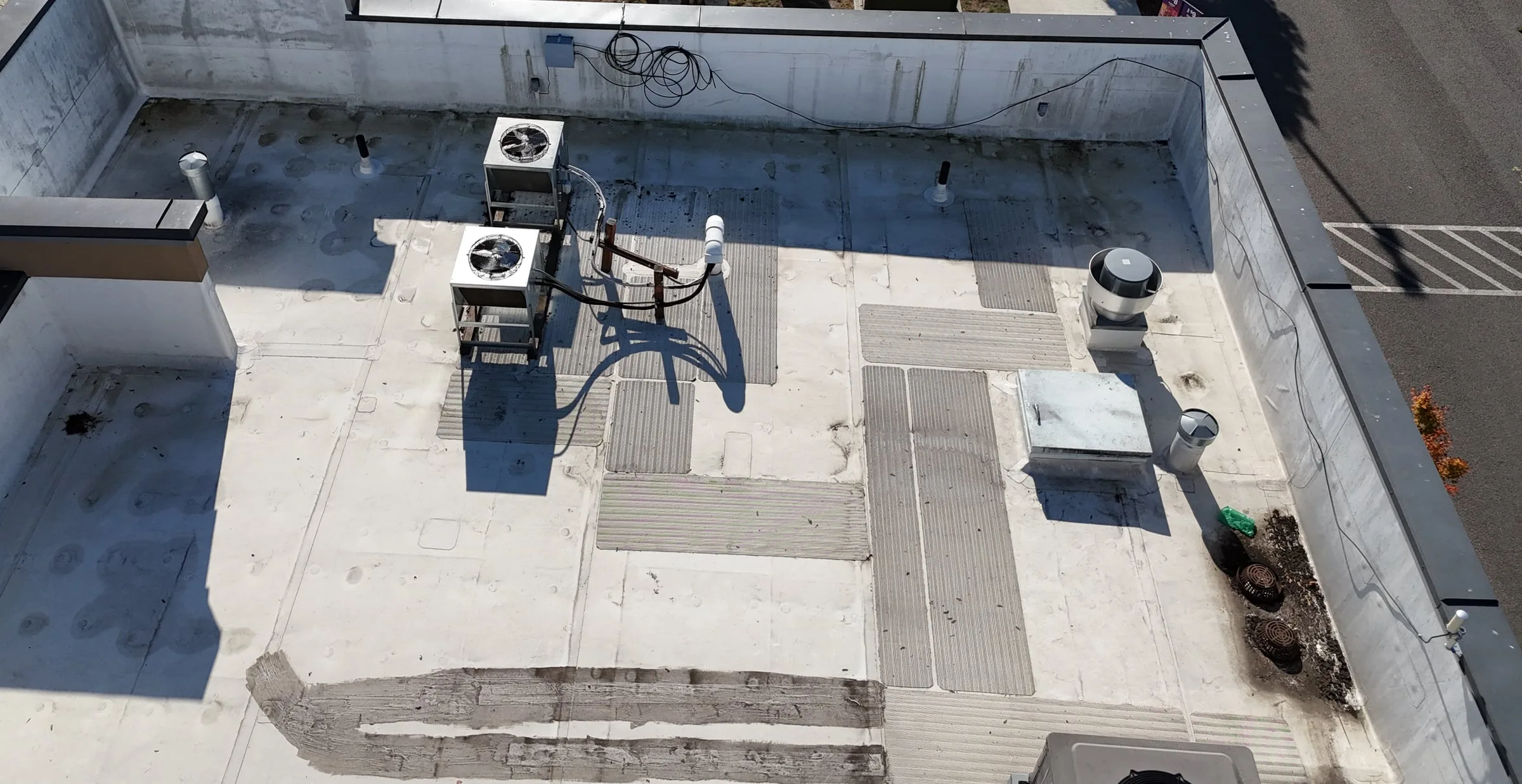 A walled flat roof from a drone, with two small AC units and their lines, and the two drains in the far corner packed with leaves and dirt.