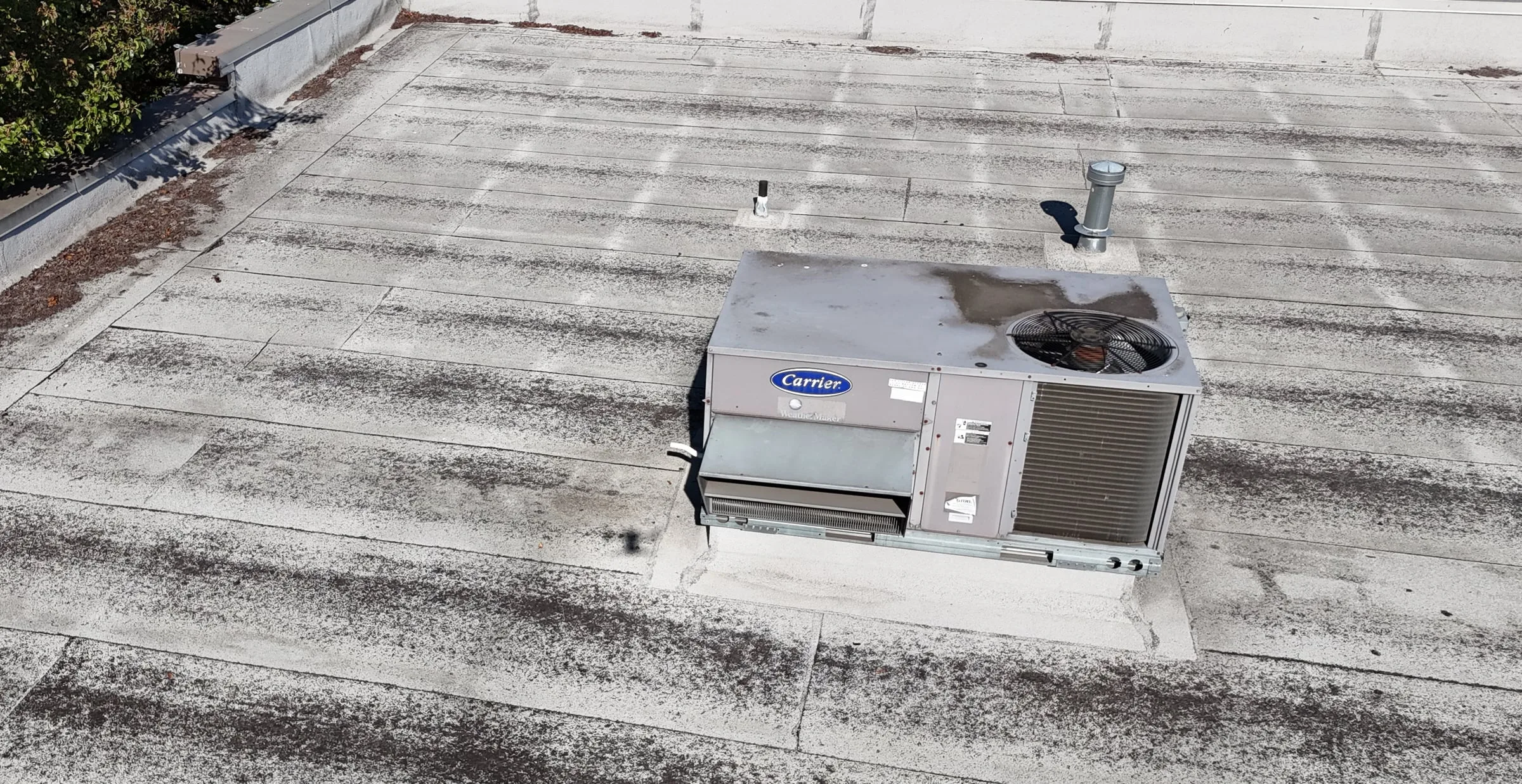 A rooftop AC unit on its curb in the middle of a weathered flat roof, with two vent pipes behind it.
