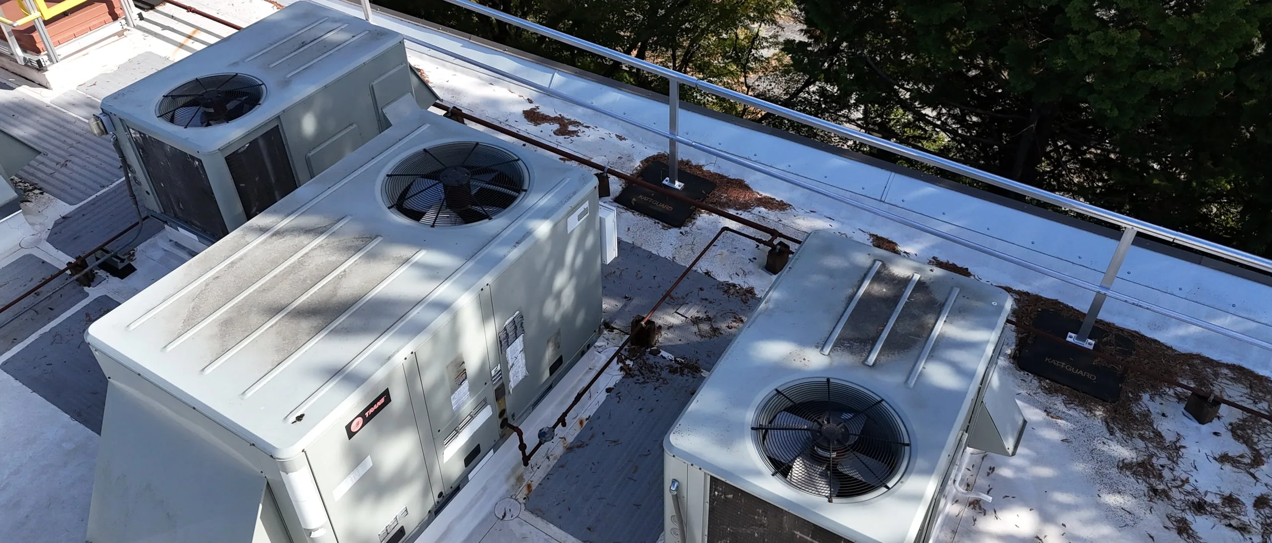 Three rooftop AC units on a white flat roof, seen from a drone, with gas lines between them, pine needles around them, and a metal safety railing.