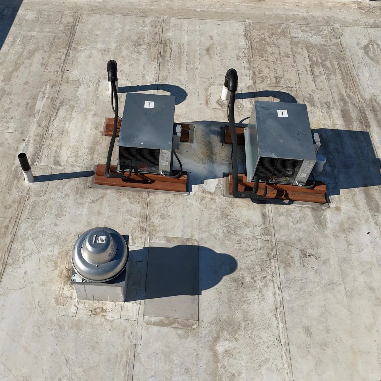 Two small AC units on wood blocks on a flat roof, their lines curving down into the roof beside them, with an exhaust fan in front.