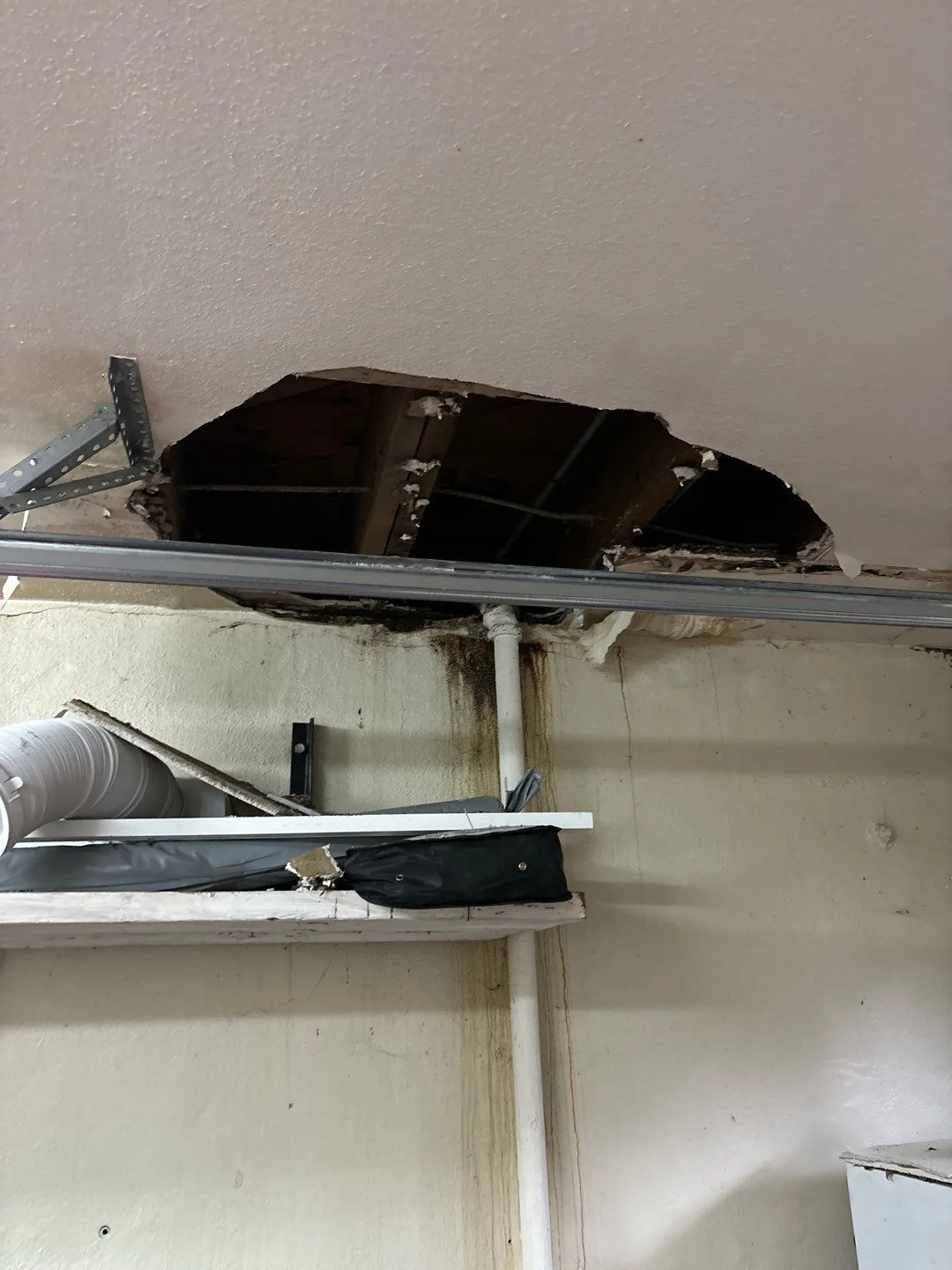A ceiling cut open to show dark, stained framing above, with brown water streaks running down the wall below it along a white pipe