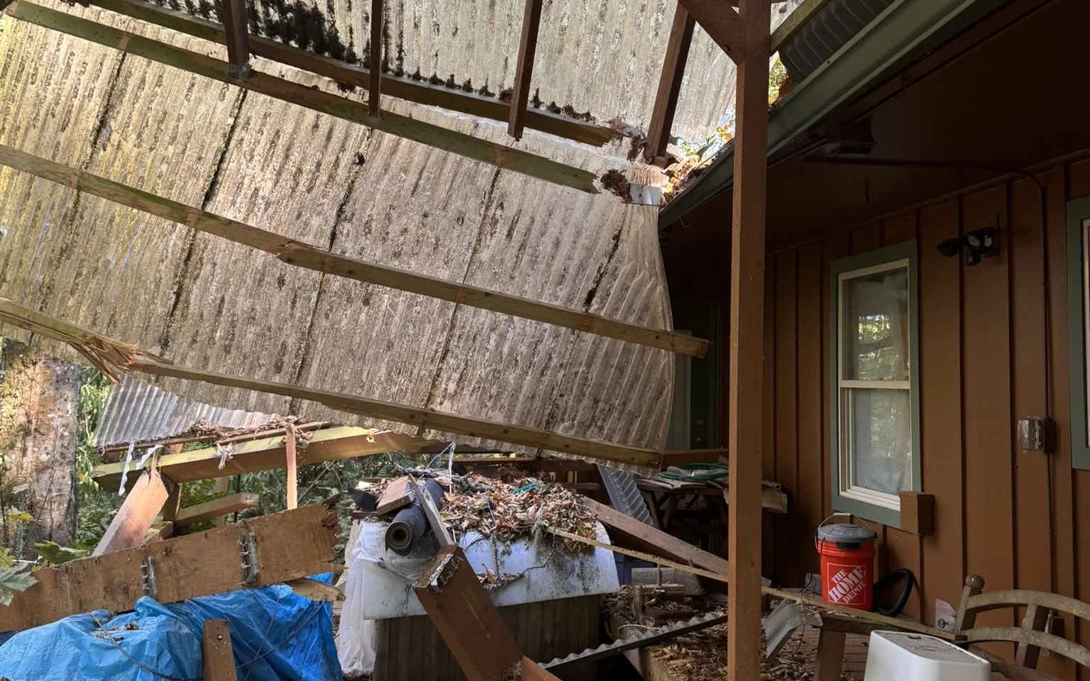 Underneath a covered patio whose corrugated panels and wood framing have dropped at a steep angle onto the deck, with debris, a blue tarp and a bin crushed beneath