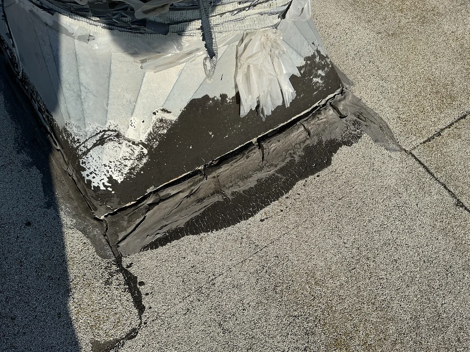 Cracked and separating waterproofing around the base of rooftop equipment beside granule-covered roofing