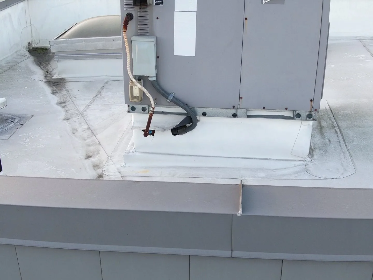 The side of a rooftop unit's curb on a white membrane roof, where the curb flashing has been cut and peeled open around a copper gas line and grey electrical conduit going into the unit