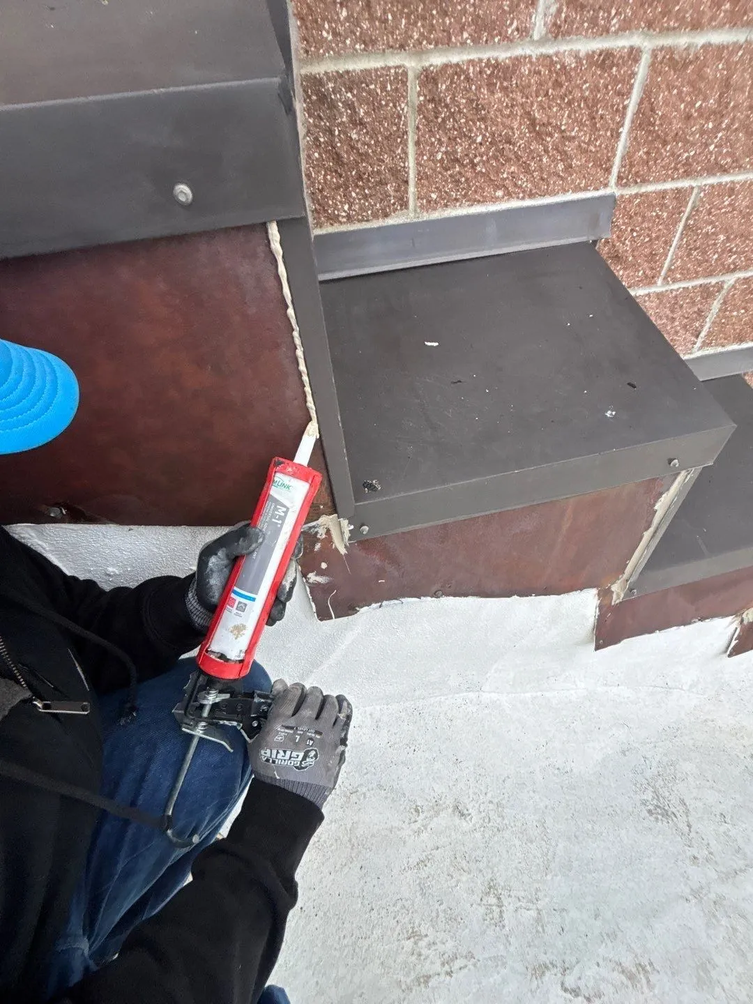 A roofer in work gloves running a bead of sealant up a vertical joint in the metal flashing on a stepped masonry wall, just above a white flat roof
