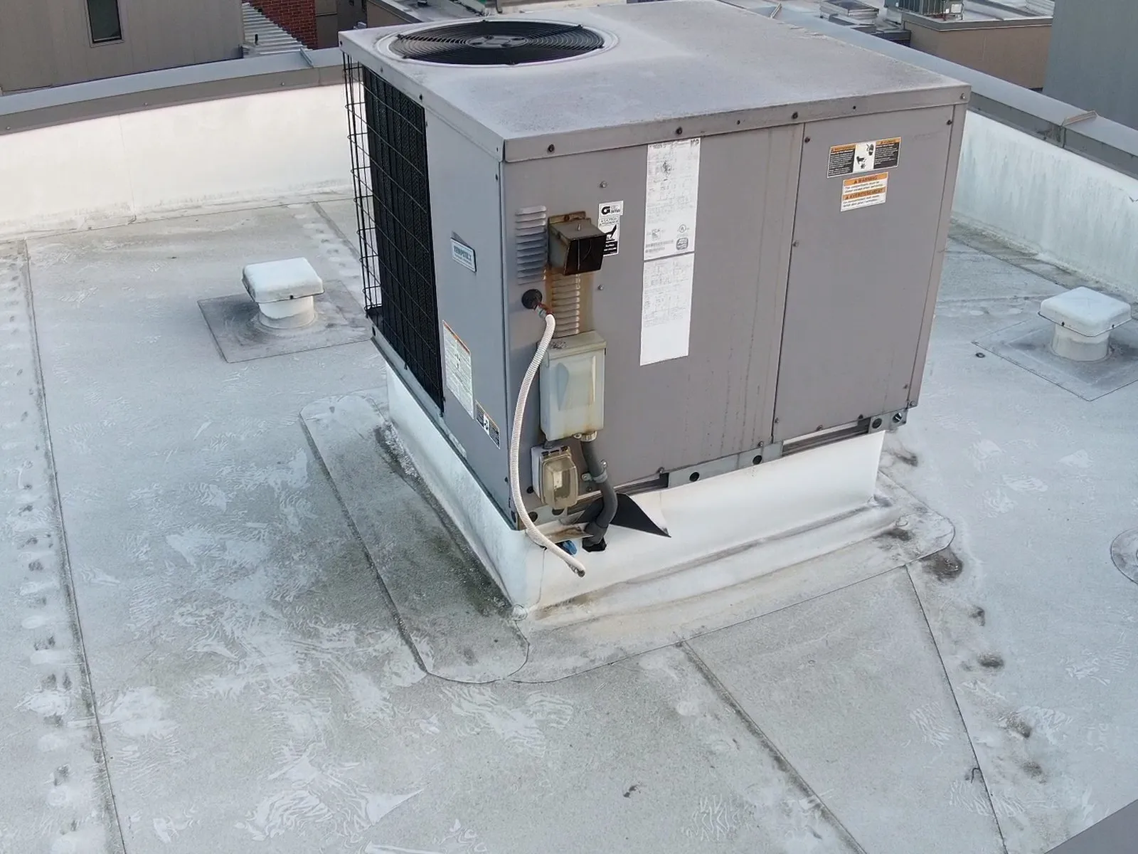 A grey packaged rooftop HVAC unit on a white single-ply flat roof, sitting on a curb wrapped in white membrane, with a gas line and electrical conduit running into the unit at one corner