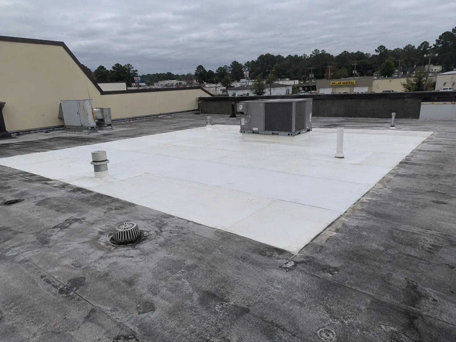 A dark, weathered commercial flat roof with a large rectangle of new white single-ply membrane in the middle, laid around a rooftop HVAC unit and several vent pipes, with the old dark roof and a drain around it