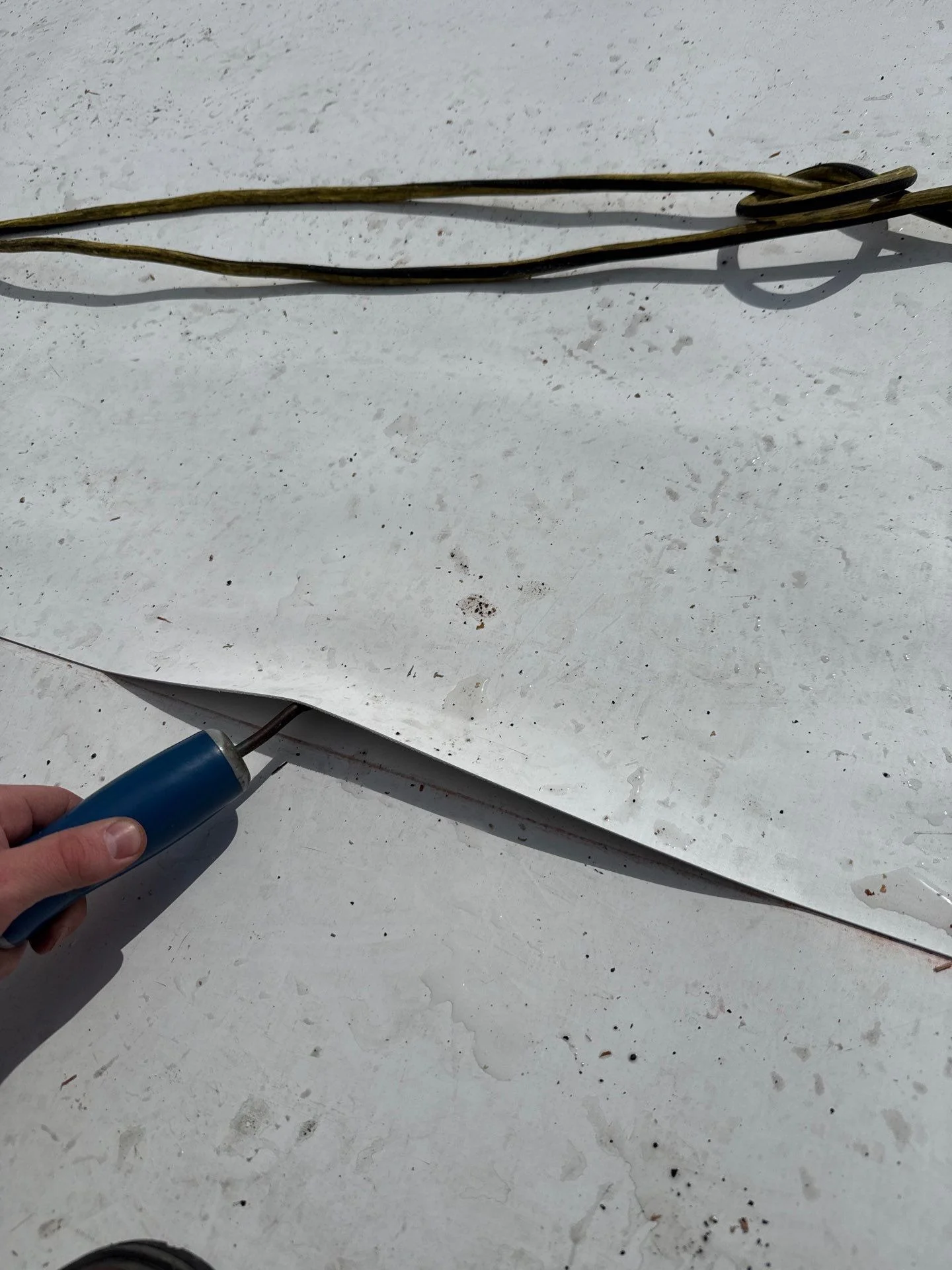 A roofer's seam probe pushed under the top sheet of a white single-ply roof seam, lifting a long stretch of the lap clear of the sheet below, with an extension cord lying on the roof behind