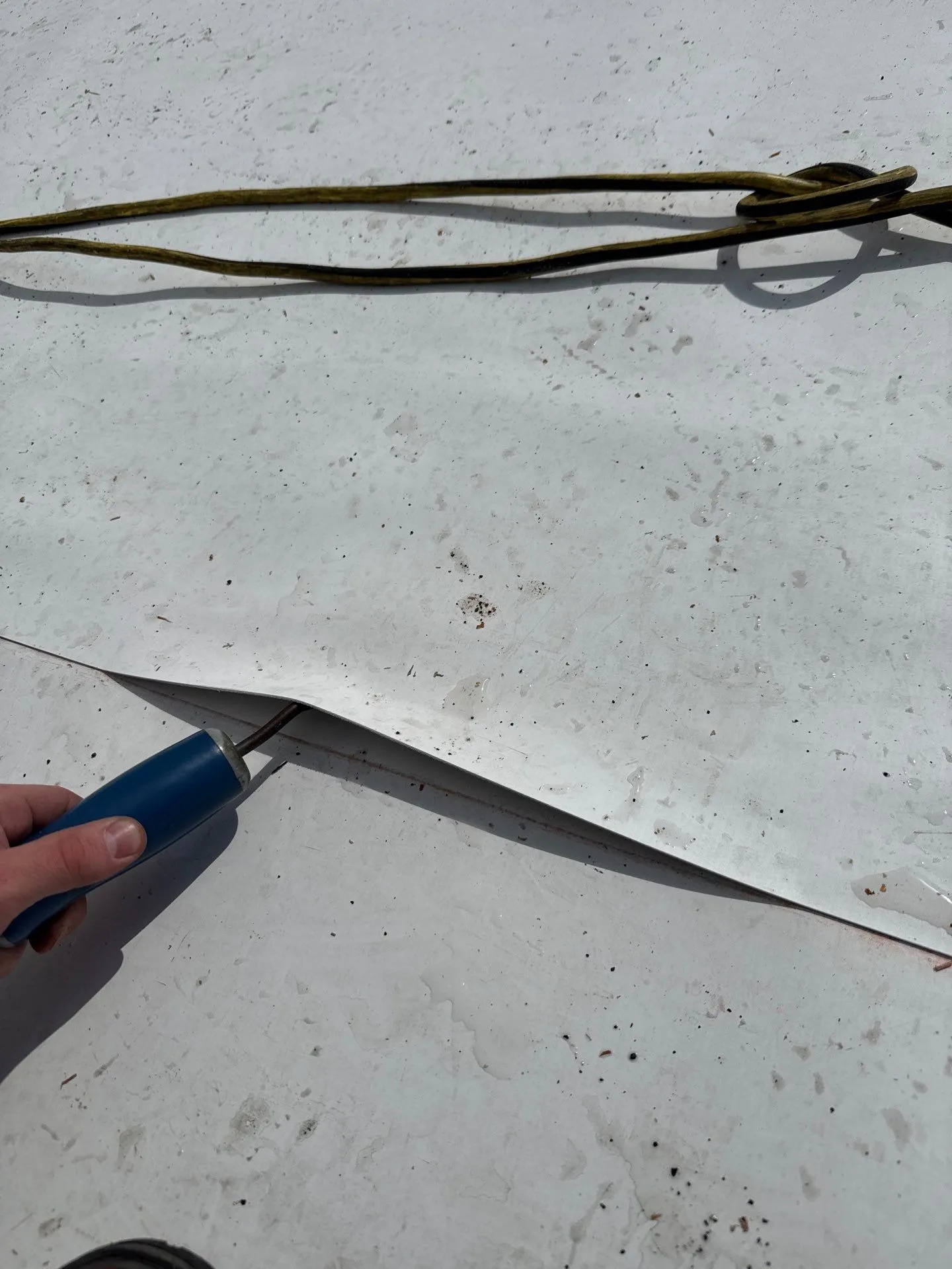 A roofer's seam probe pushed under the top sheet of a white single-ply roof seam, lifting a long stretch of the lap clear of the sheet below, with an extension cord lying on the roof behind