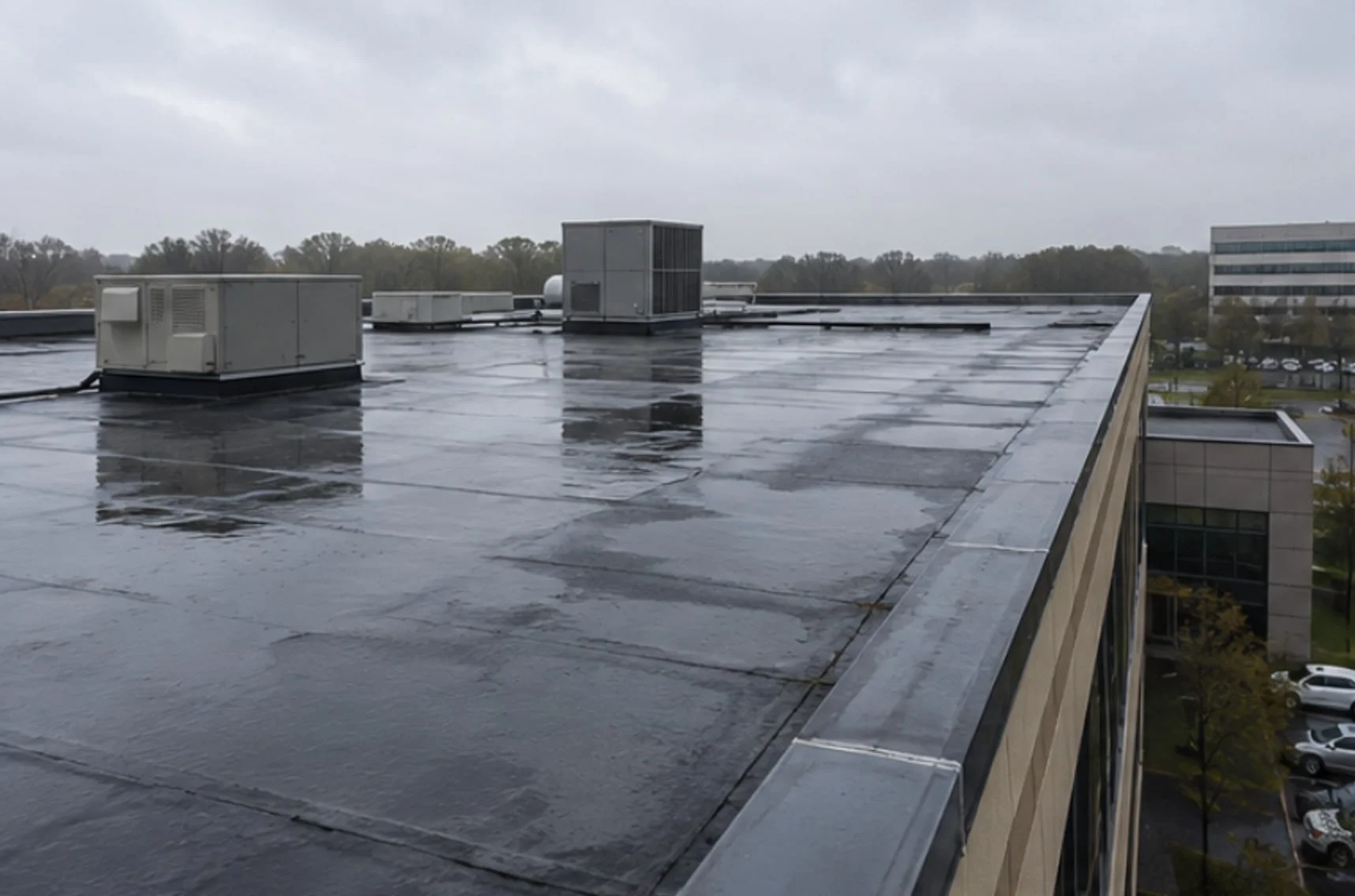 A wet black flat roof on a commercial building under an overcast sky, with seams running across the membrane, two rooftop units near the far edge, and a metal-capped parapet along the side