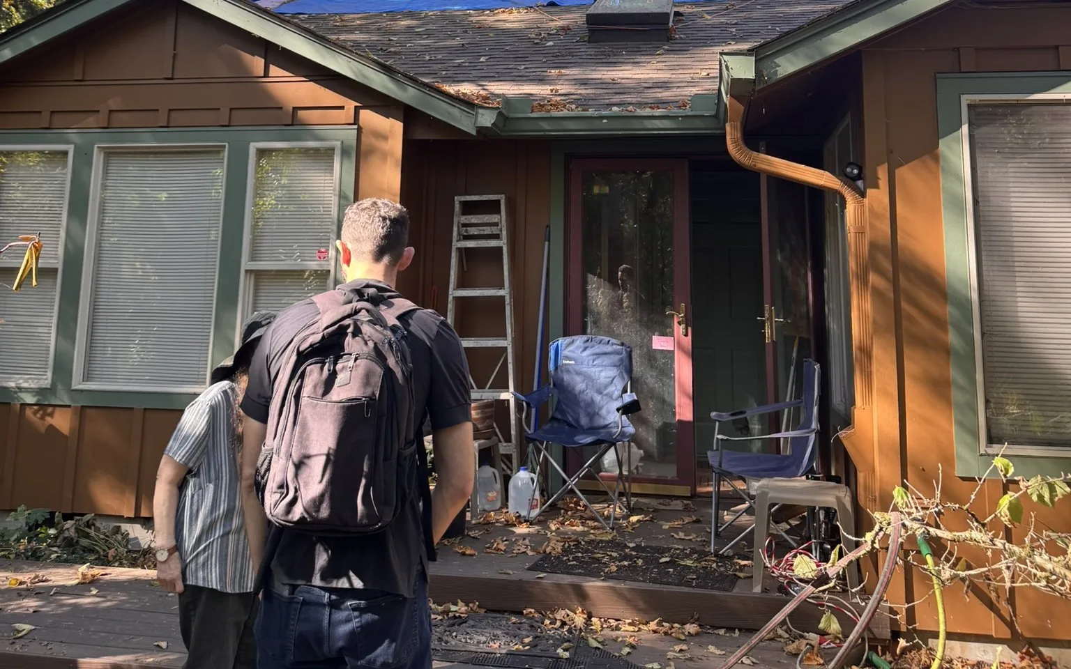 Two people seen from behind on the front deck of a wood-sided house, a man with a backpack and a person in a striped shirt, facing the front door with a ladder and folding chairs beside it
