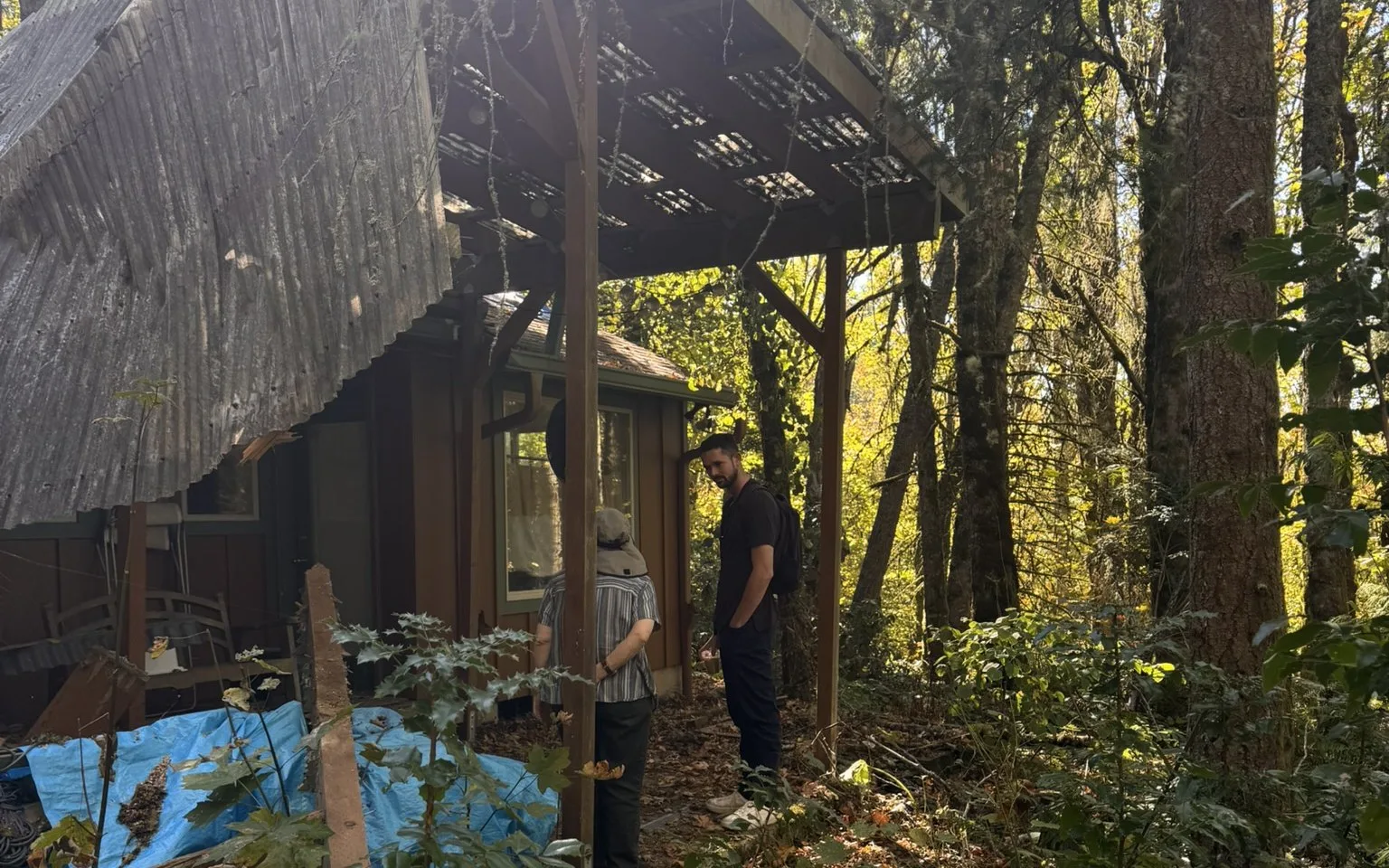 A man in a black shirt and backpack, and a person in a striped shirt and hat seen from behind, standing and talking under the part of a patio frame that is still upright beside the house, with a sagging corrugated panel overhead and tall trees behind