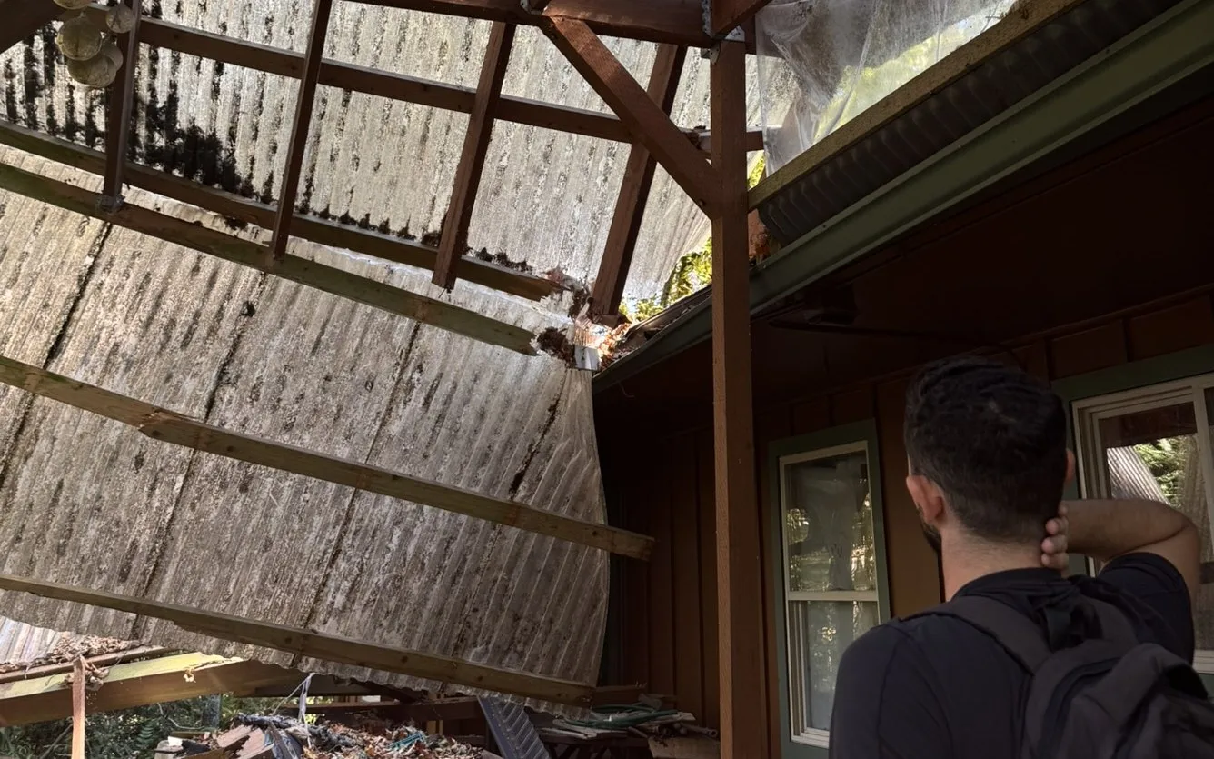 A man seen from behind, hand at the back of his head, looking up at a patio cover whose wood framing and corrugated panels sag down at a steep angle beside the house