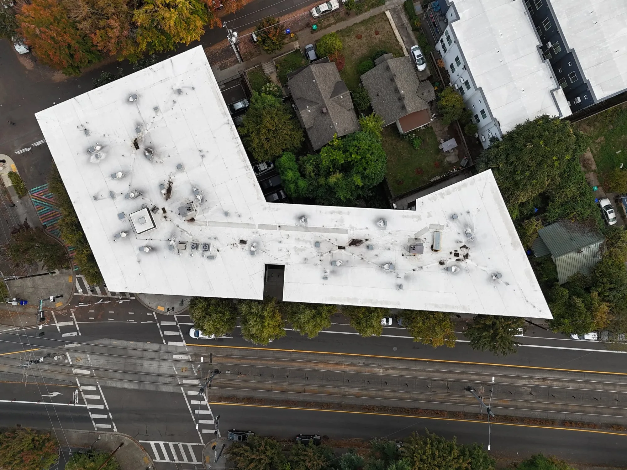 A large white flat roof on an L-shaped building, seen from straight above, dotted with vents and patched spots.