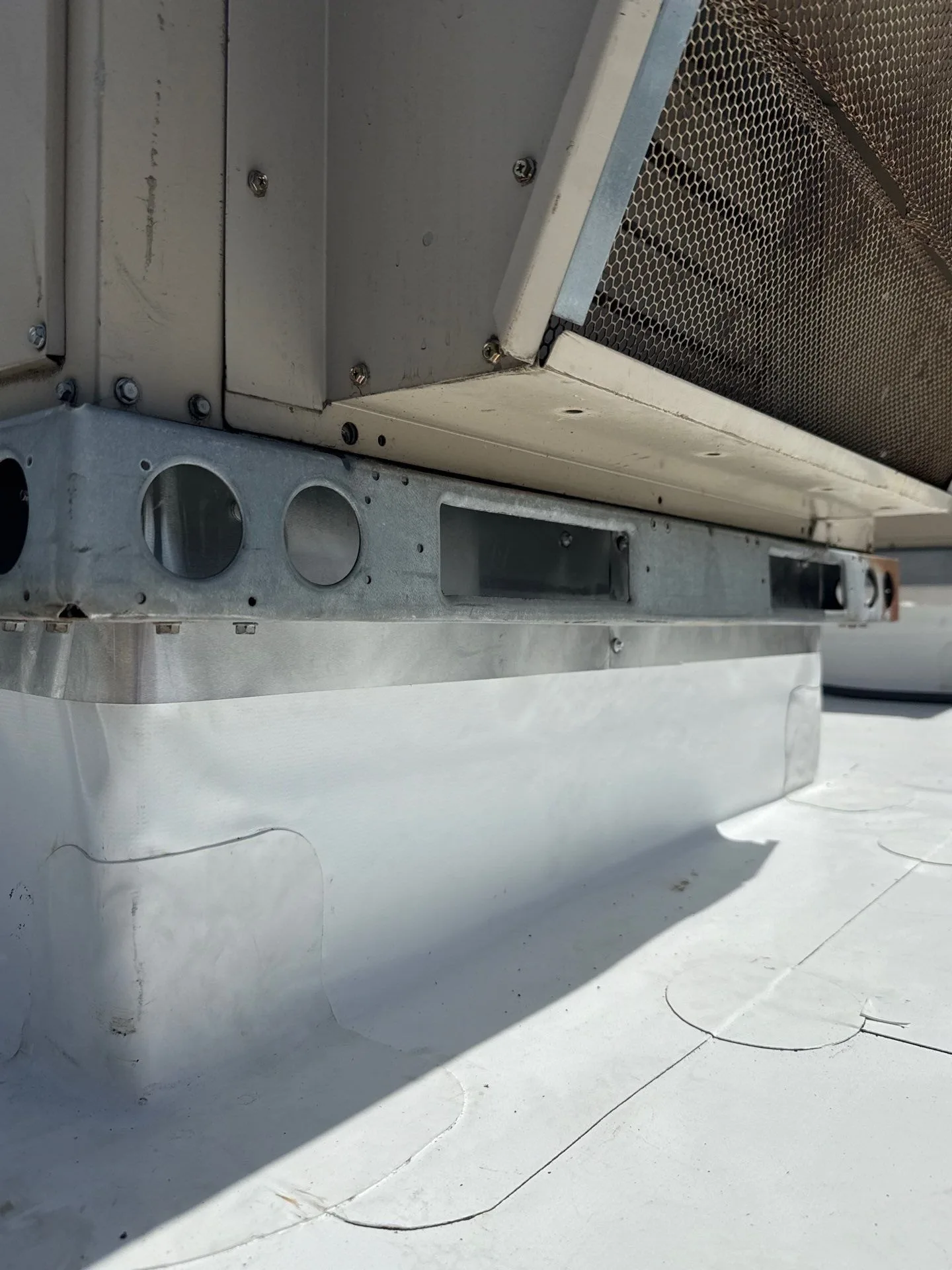 The base of a rooftop HVAC unit on a white single-ply roof, with the membrane running up the sides of the curb, rounded patches welded over the corners, and a strip of metal flashing along the top edge under the unit's steel base rail