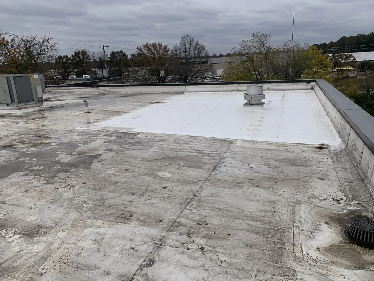 An older light gray single-ply flat roof with a bright white new section around an exhaust fan, running to the parapet, and a roof drain in the corner of the old roof just outside the new section