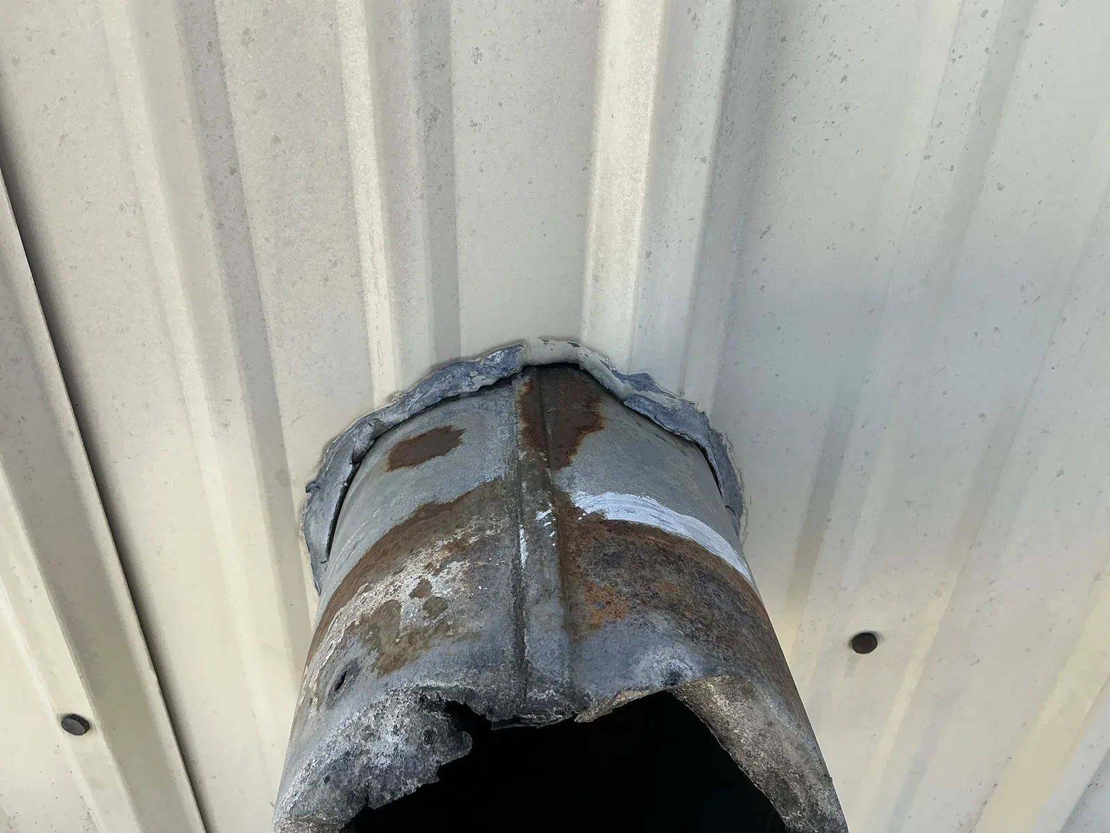 Close view of separated sealant around a corroded pipe passing through a ribbed metal wall.