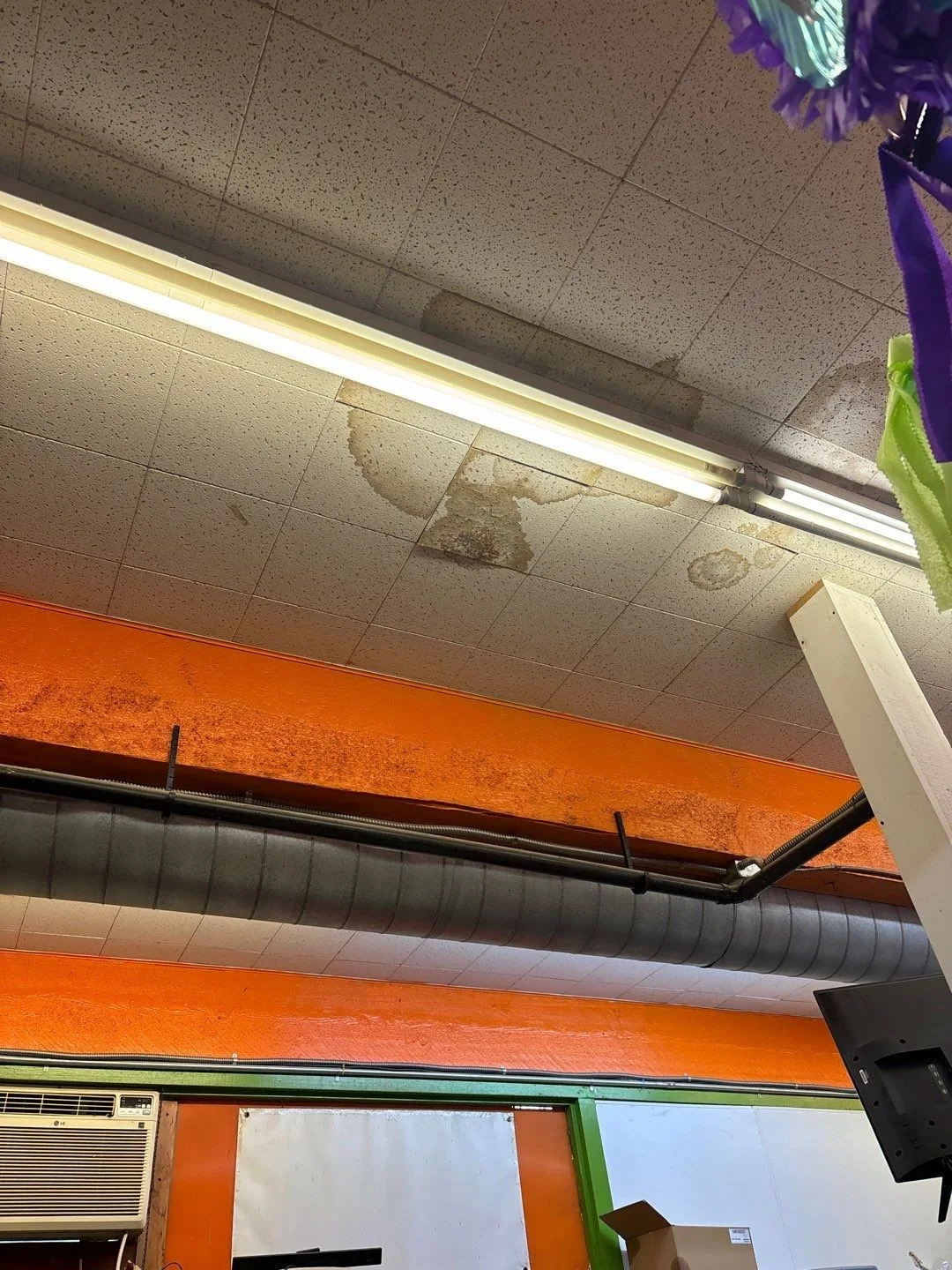 Ceiling tiles along a fluorescent light fixture with several brown water stains, some tiles showing rings inside rings, above an orange beam, a black duct and a wall-mounted air conditioner