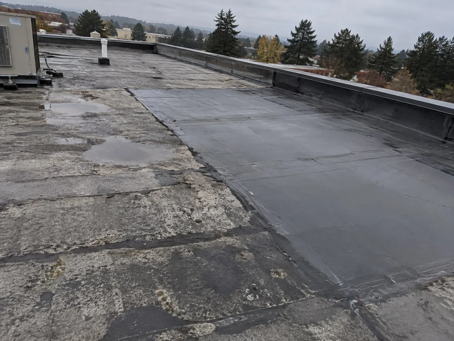 A weathered gray asphalt flat roof with puddles of standing water on the left, and a large section of new dark roofing on the right running along the parapet, joined to the old roof along a black sealed edge