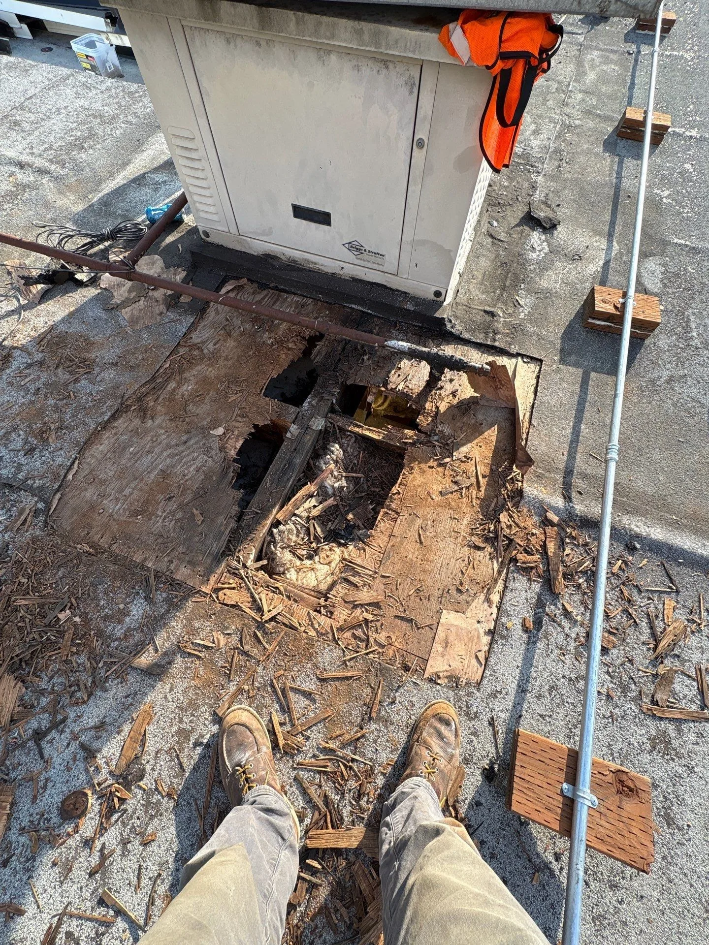 A section of flat roof cut open beside a rooftop generator, showing plywood decking rotted through to the joists, broken wood scattered around the hole and old insulation visible inside