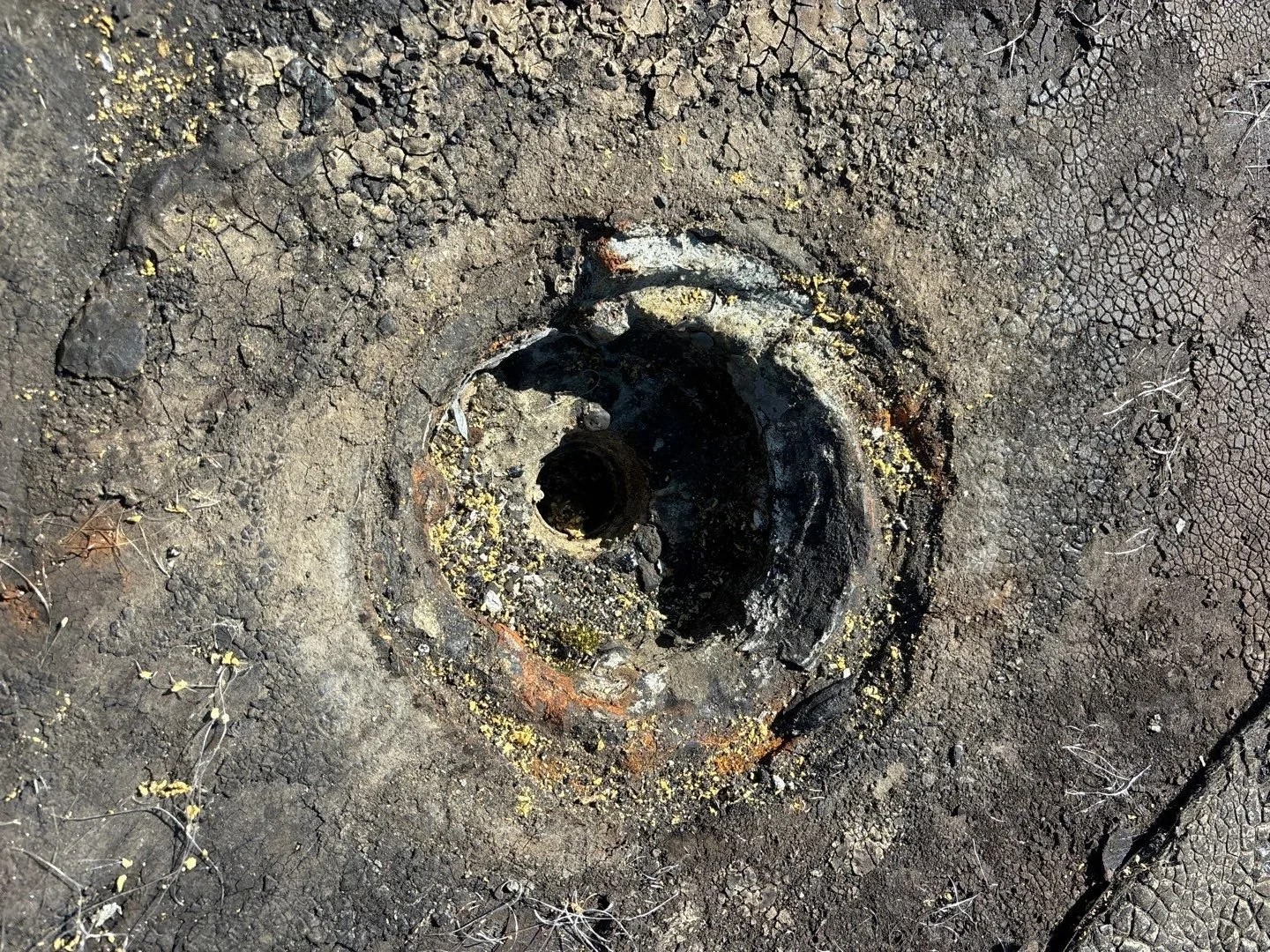 An old cast-iron roof drain with no strainer, its rim rusted orange, and the cracked, weathered roofing around it broken away from the edge of the bowl, with moss and debris inside
