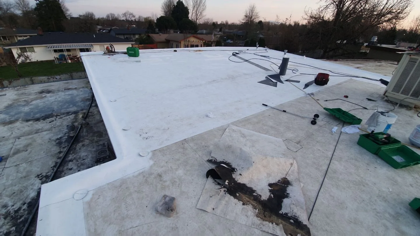 New white membrane being installed across part of the same flat roof, with welding equipment nearby and a cut-out piece of the old roofing in front with black mastic still stuck to it