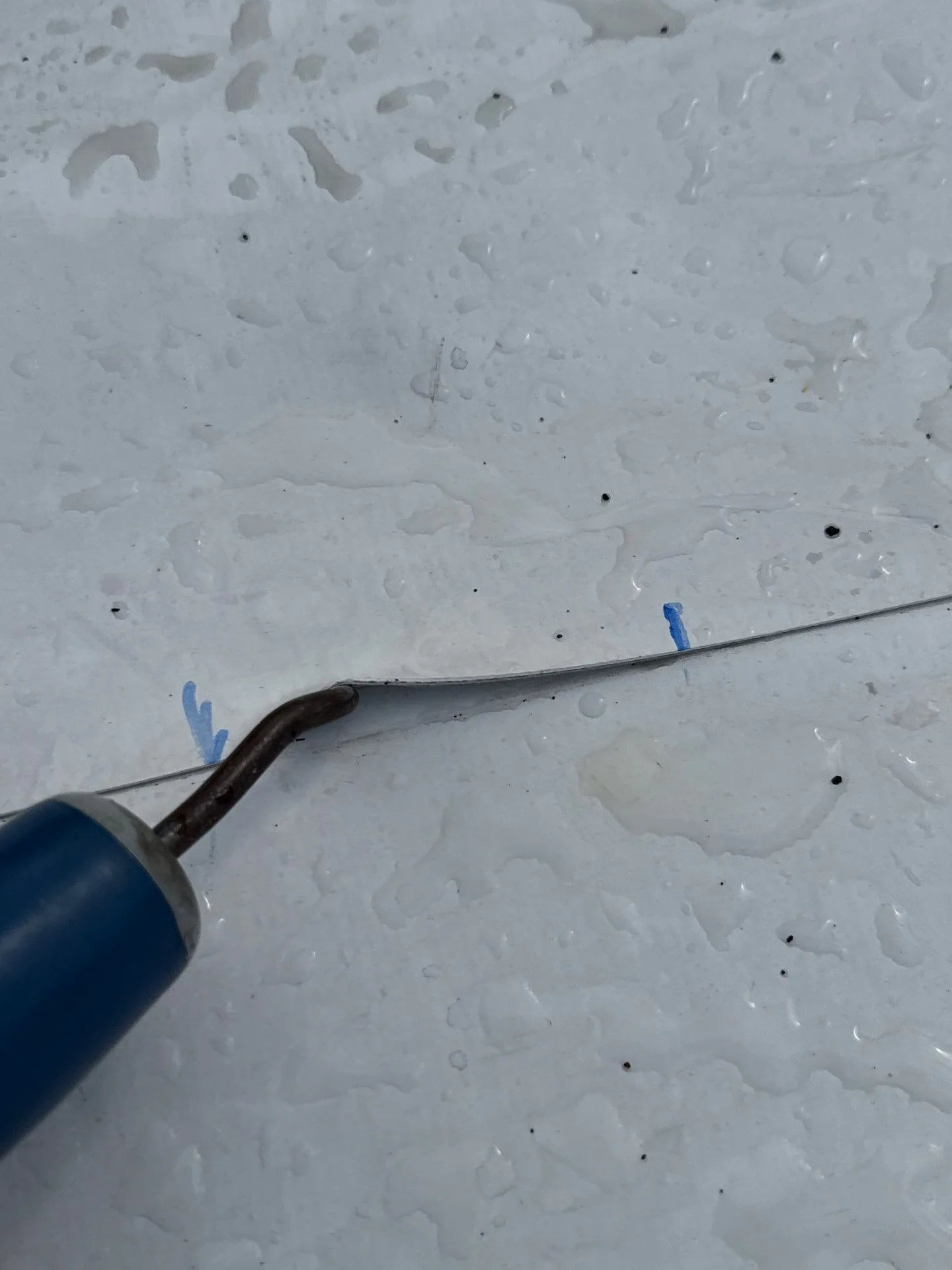 Close-up of a seam probe hooked under the edge of a white single-ply roof seam, with two blue crayon marks on the membrane on either side of the open stretch