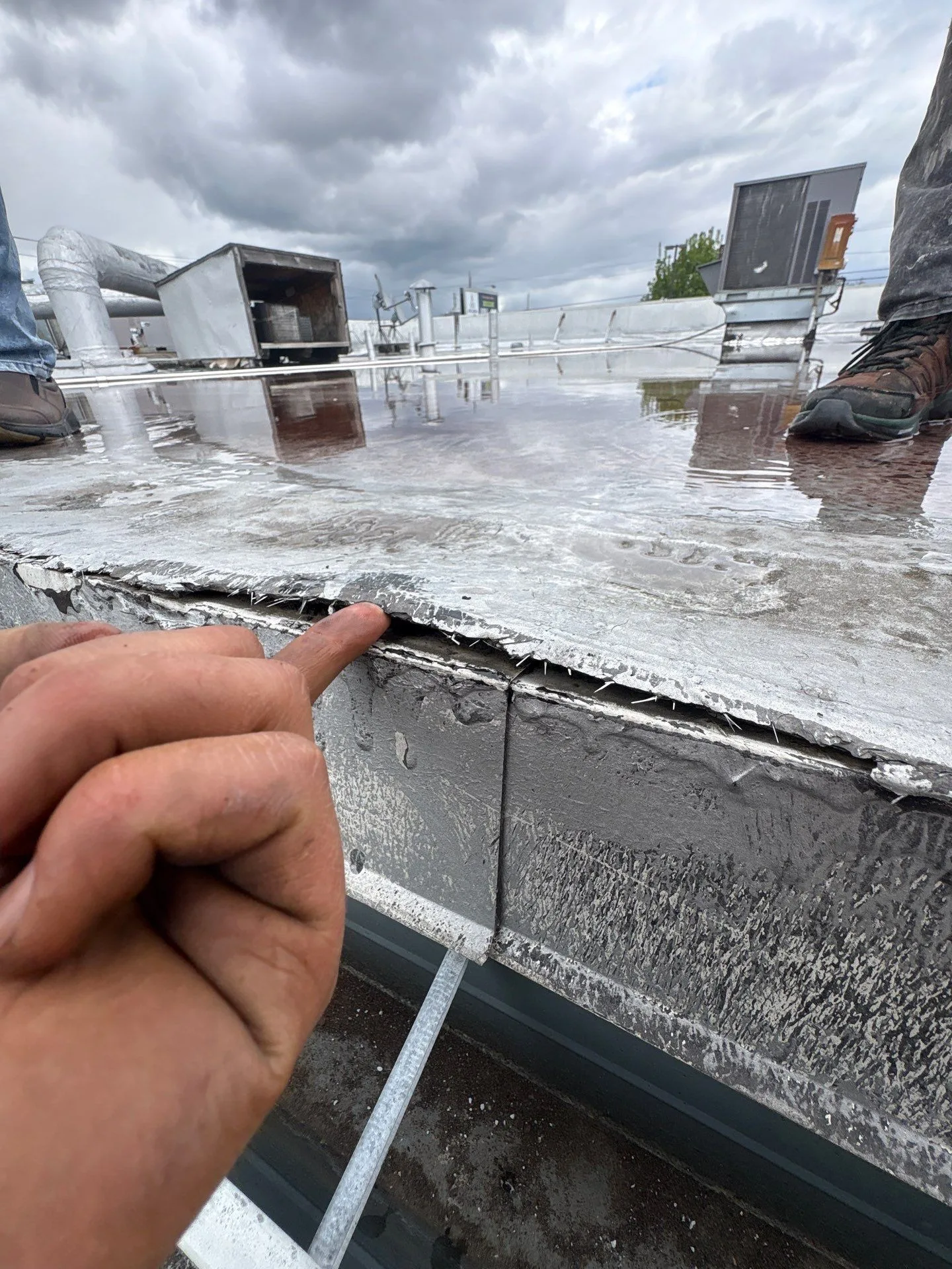 A finger pointing into a gap where silver-coated roofing has lifted off the metal edge of a wet flat roof, with the dark joint between two pieces of edge metal visible below