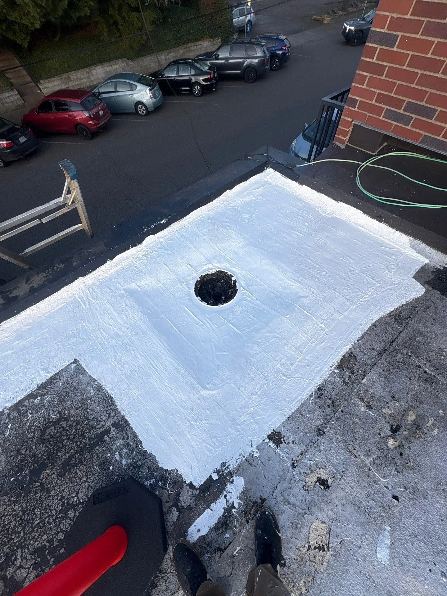 A wide area of fresh white silicone around a roof drain on an older dark flat roof beside a brick wall, with a parking lot below