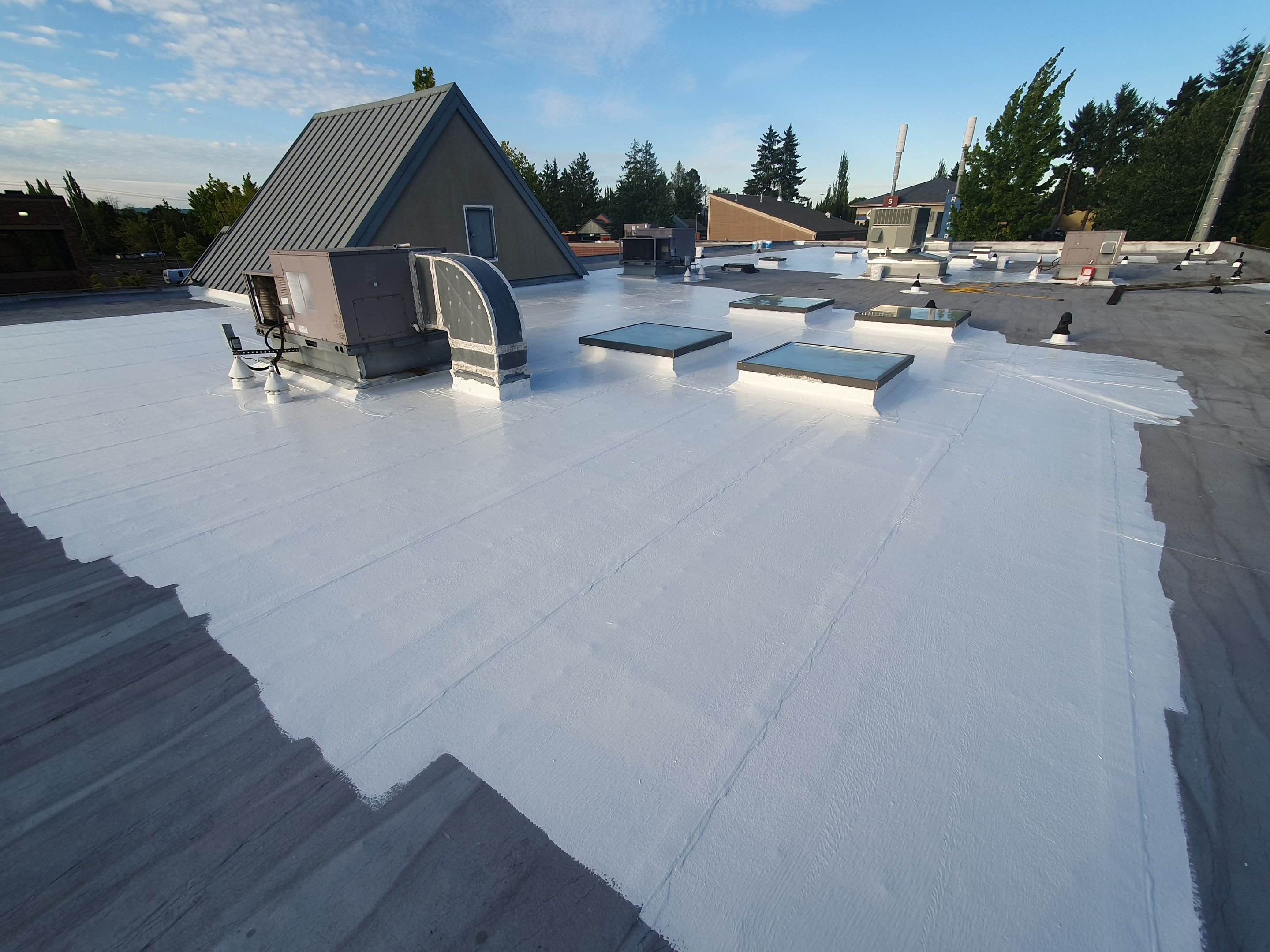 A commercial flat roof mid-reseal, white silicone covering most of the field with the grey existing membrane still visible at the edge