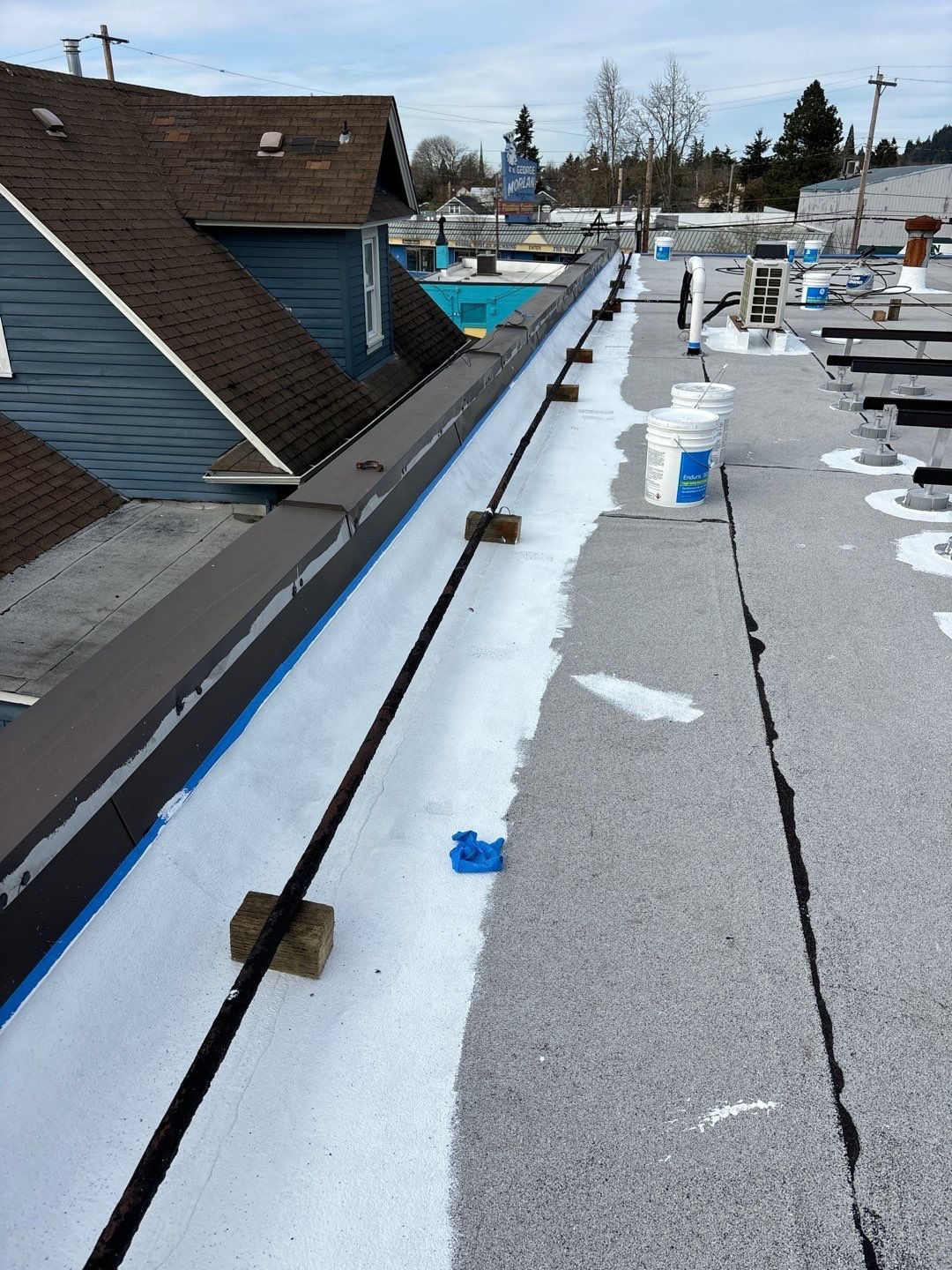 Fresh white silicone applied along the base of a parapet wall and up the wall over a blue tape line, with GE Enduris pails on the roof