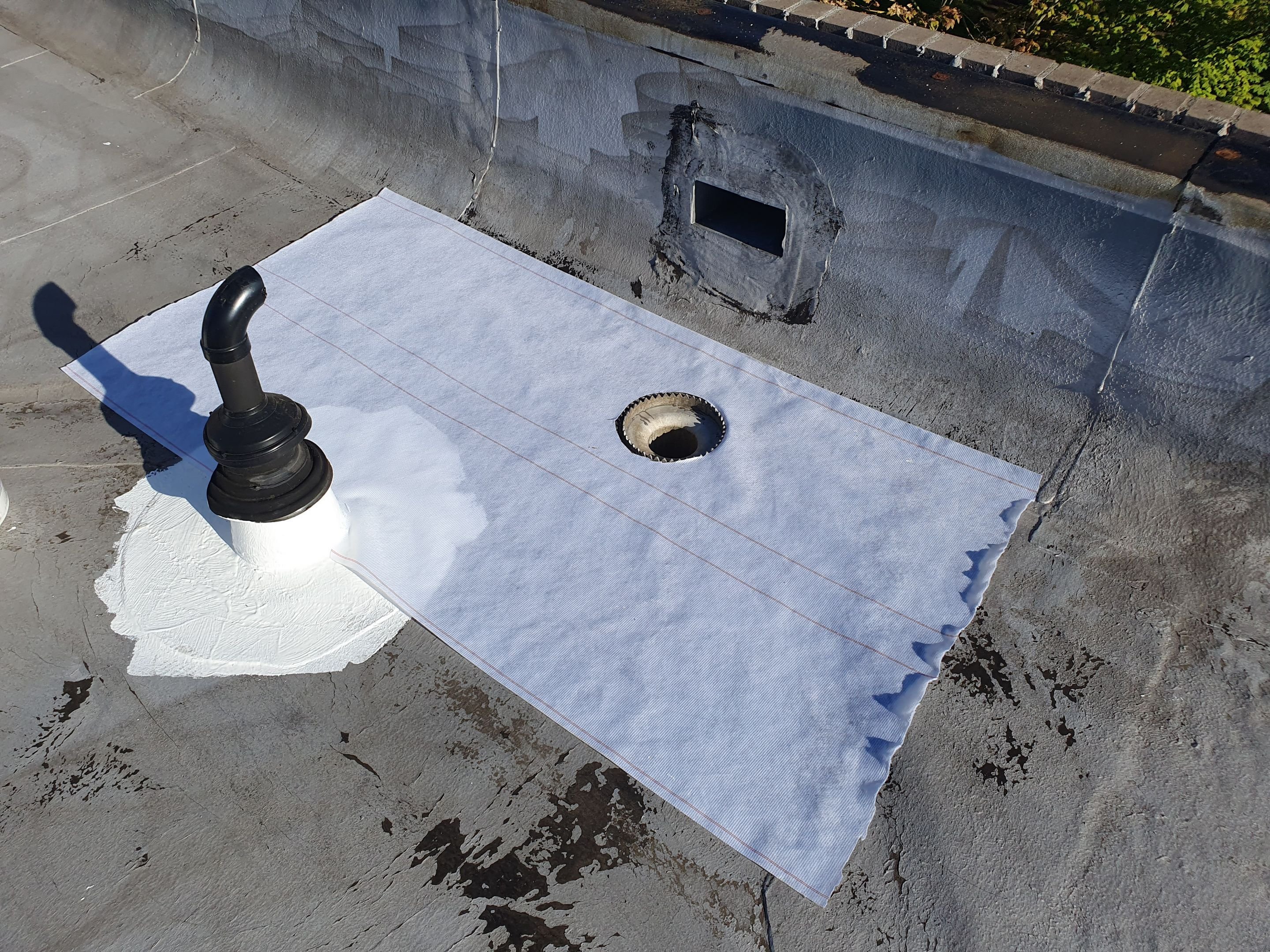 An aged flat roof with reinforcing fabric laid over a drain area and a sealed pipe boot, repaired before the silicone coat goes on
