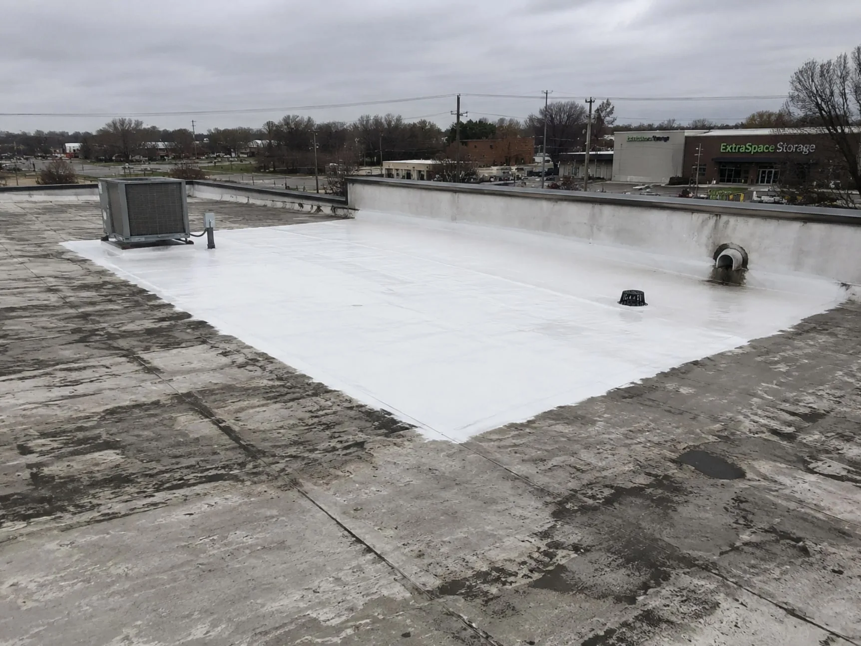 A weathered gray flat roof with one area coated bright white in silicone, running from the parapet wall out past a rooftop HVAC unit, with a drain and a wall scupper inside the coated area