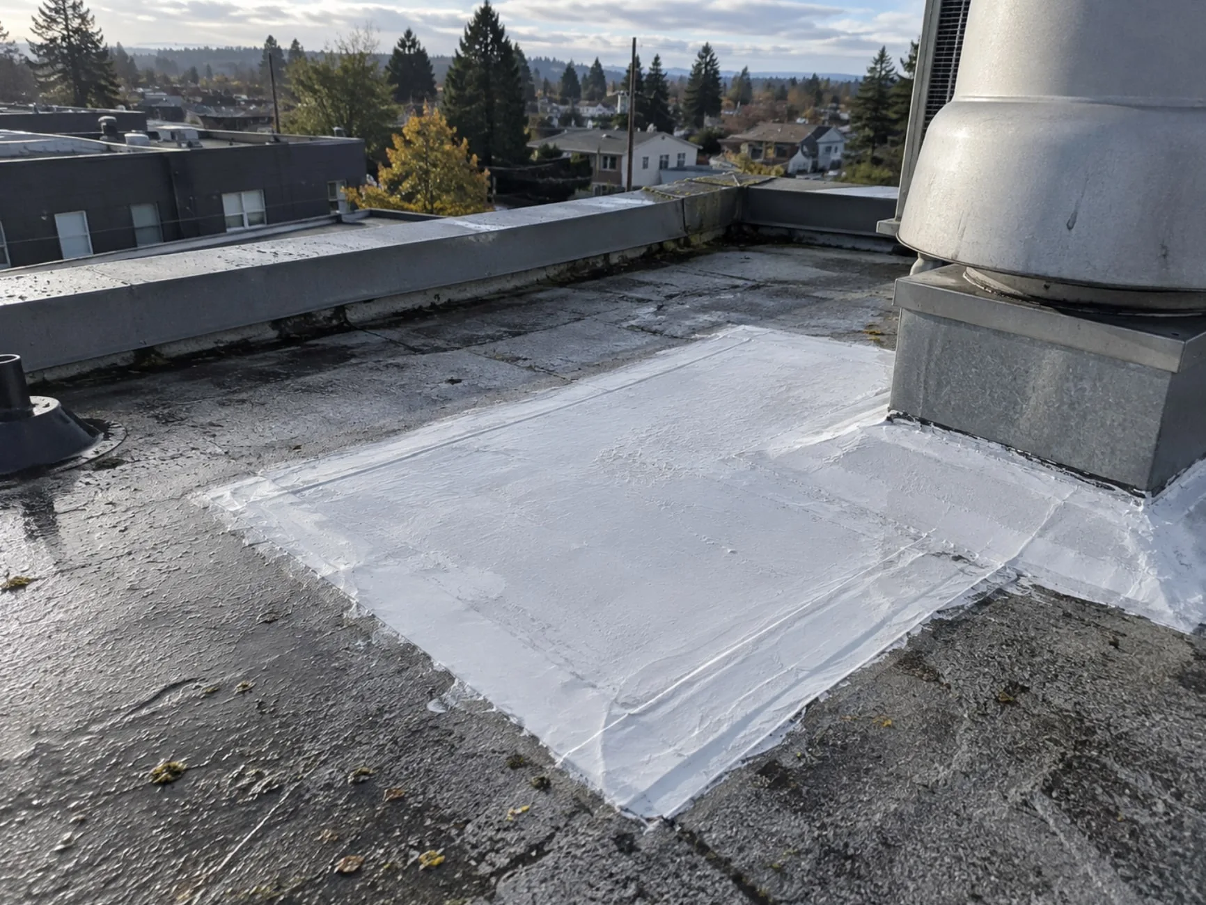 A white silicone-coated area on a dark granulated flat roof next to a large metal exhaust fan curb, with the coating carried up the side of the curb