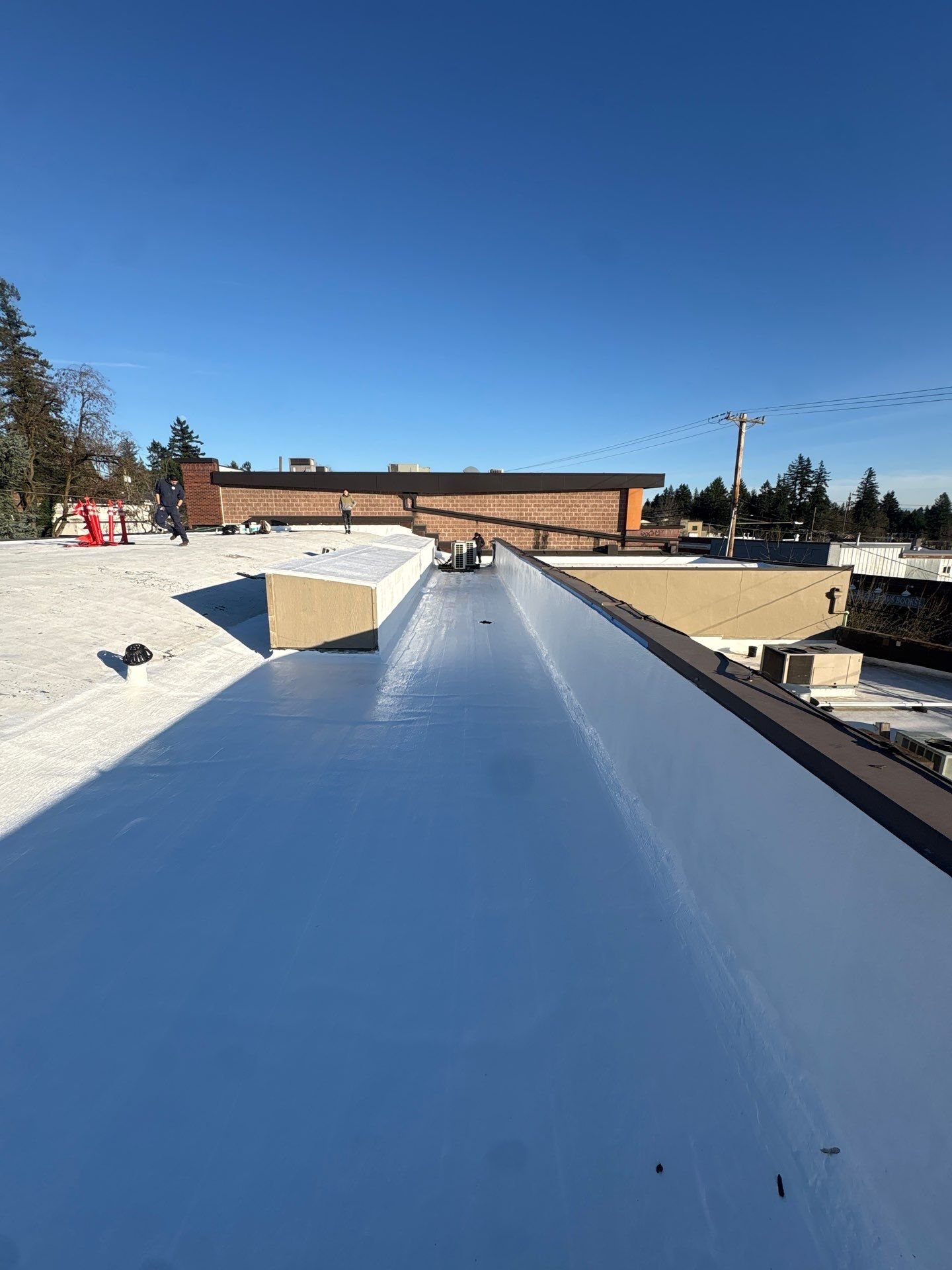 A freshly coated white roof running toward a brick parapet under a deep blue sky, with two crew members far in the distance