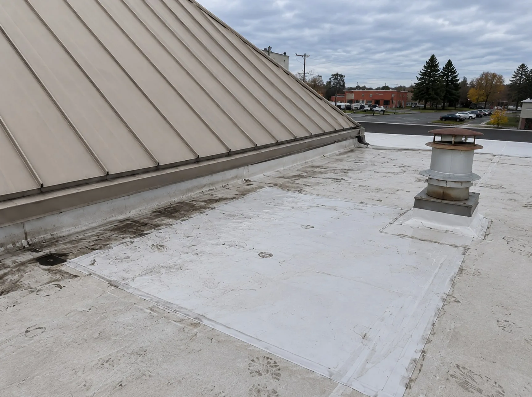 A newer white membrane section on an older white single-ply roof, running from the base of a standing-seam metal roof slope out to a vent stack, with the vent curb freshly flashed