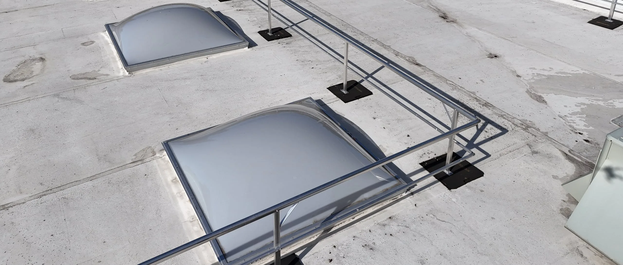 Dome skylights on a white flat roof, seen from a drone, beside a metal safety railing.