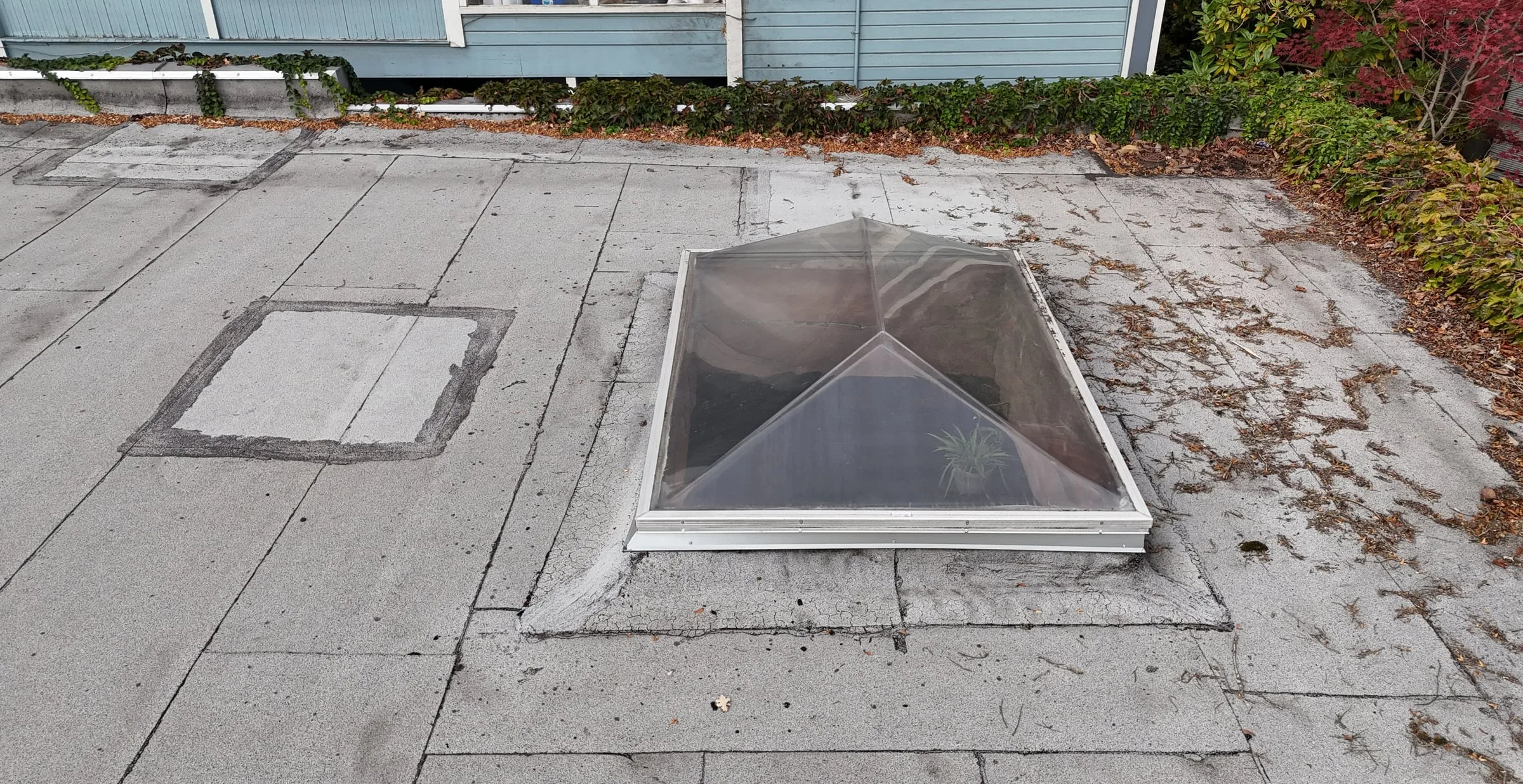 An older glass pyramid skylight on a weathered flat roof, beside a house wall.