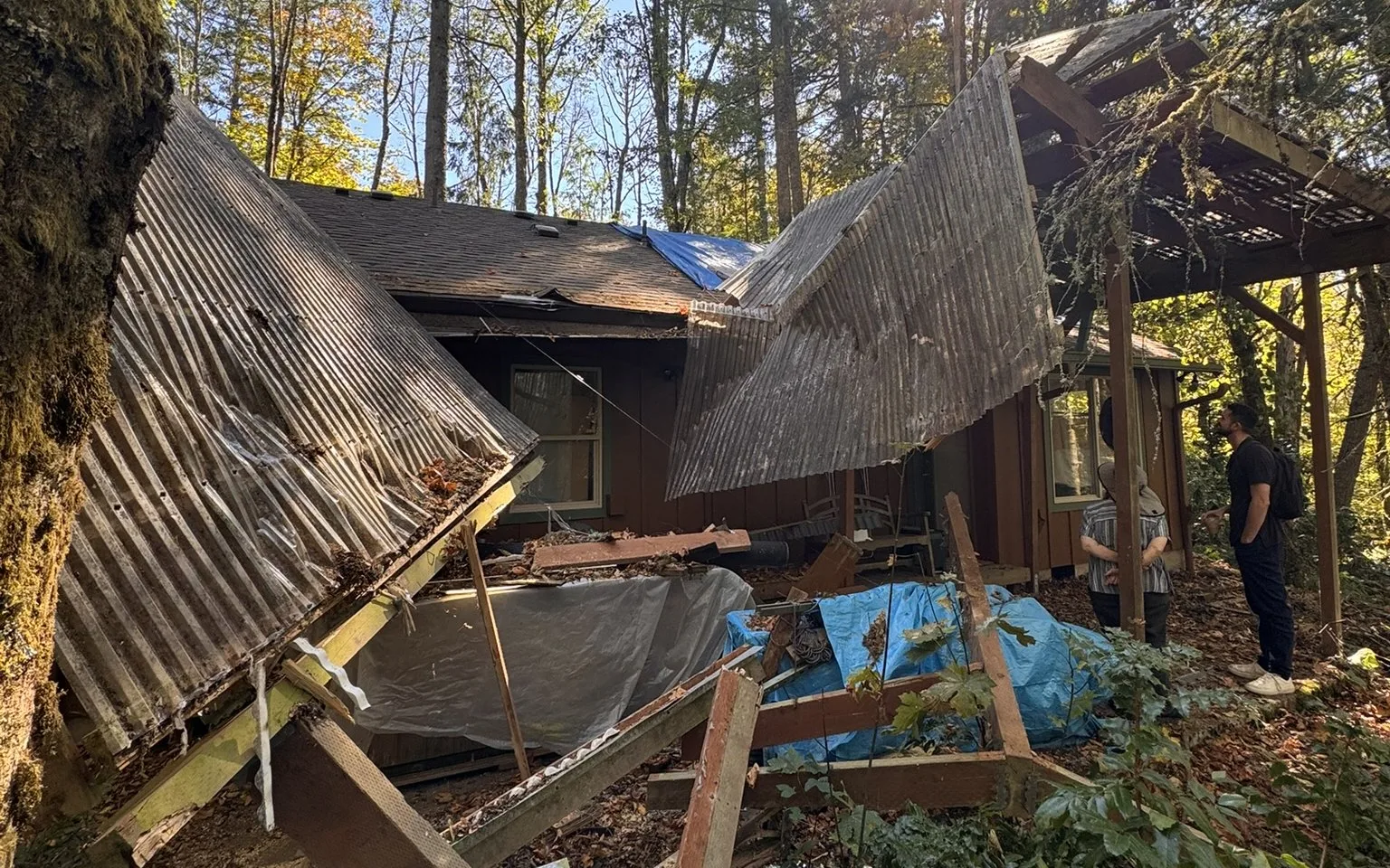 Two collapsed corrugated patio-cover panels folded down over a broken deck behind a wood-sided house, a blue tarp on the shingle roof beyond them, and two people standing under the remaining patio framing at right