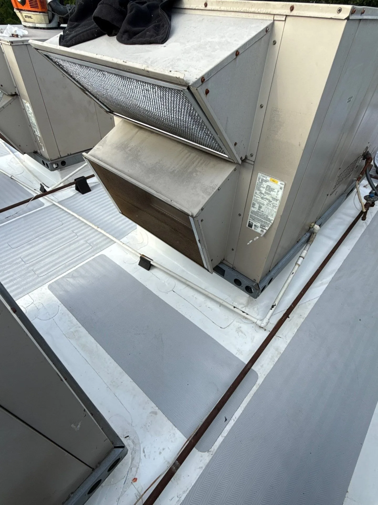 A white silicone-coated flat roof around a rooftop HVAC unit, with grey textured walkway pads laid along both sides of the unit and a condensate line and gas pipe running across the roof on supports