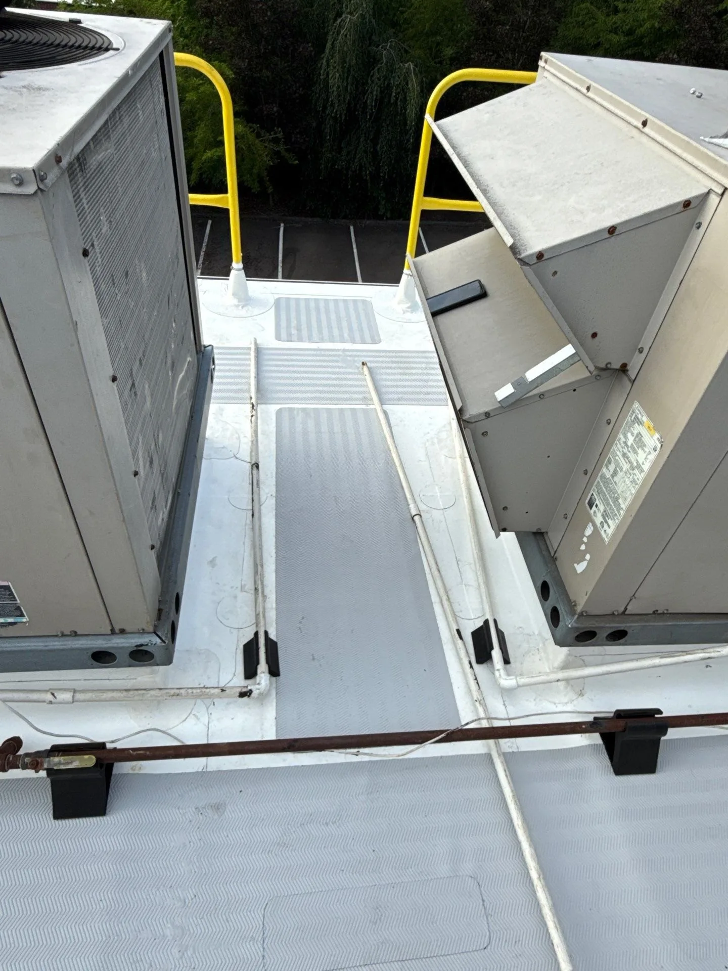 A grey textured walkway path running between two rooftop HVAC units on a white roof, toward a yellow safety railing at the roof edge
