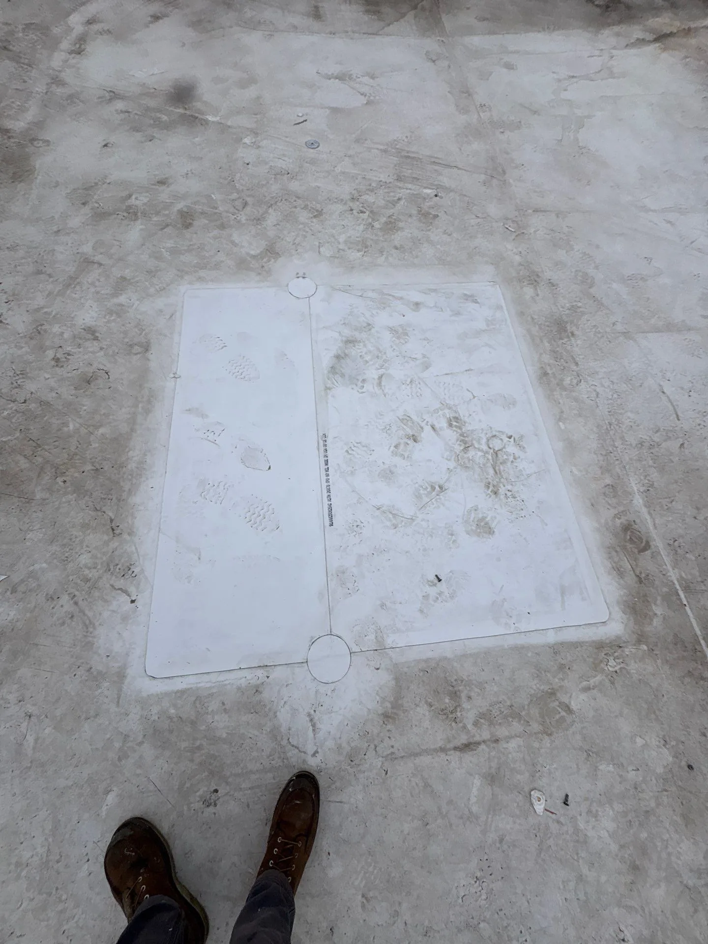 A clean white rectangular patch welded onto a dirty white flat roof, with a seam down its middle and a round cover piece at each end of the seam, a roofer's boots at the bottom of the frame